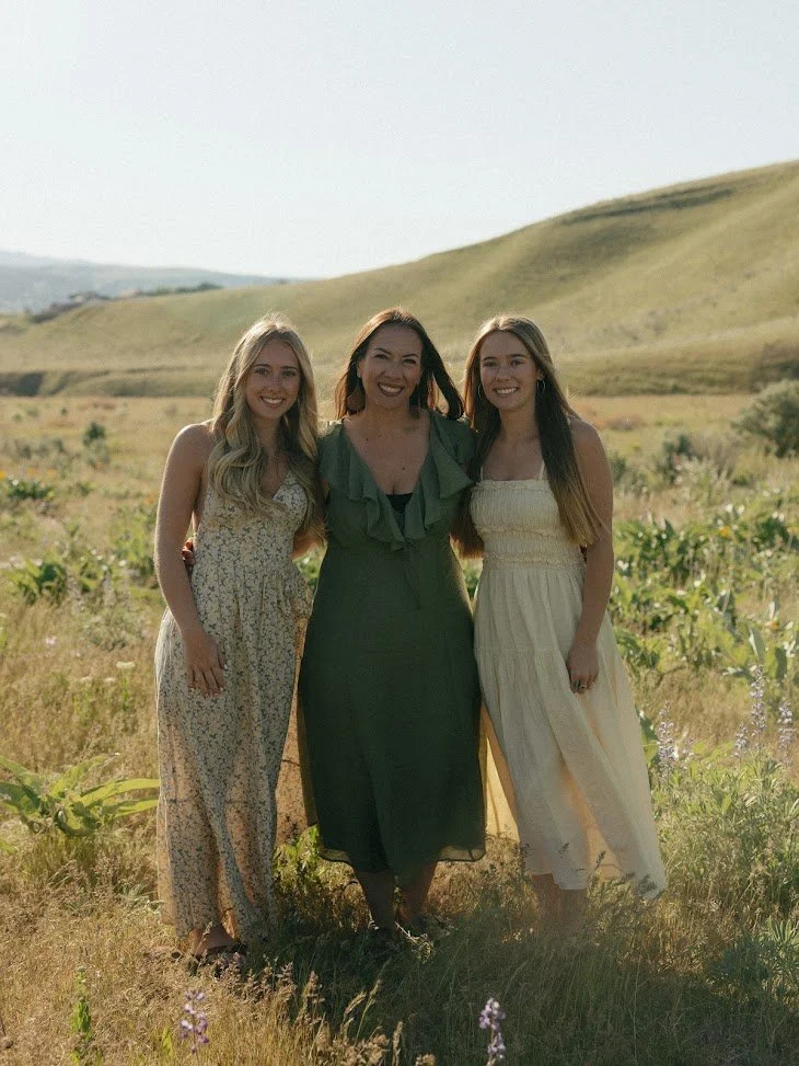 Three women standing outdoors in a grassy field with rolling hills in the background, smiling at the camera.