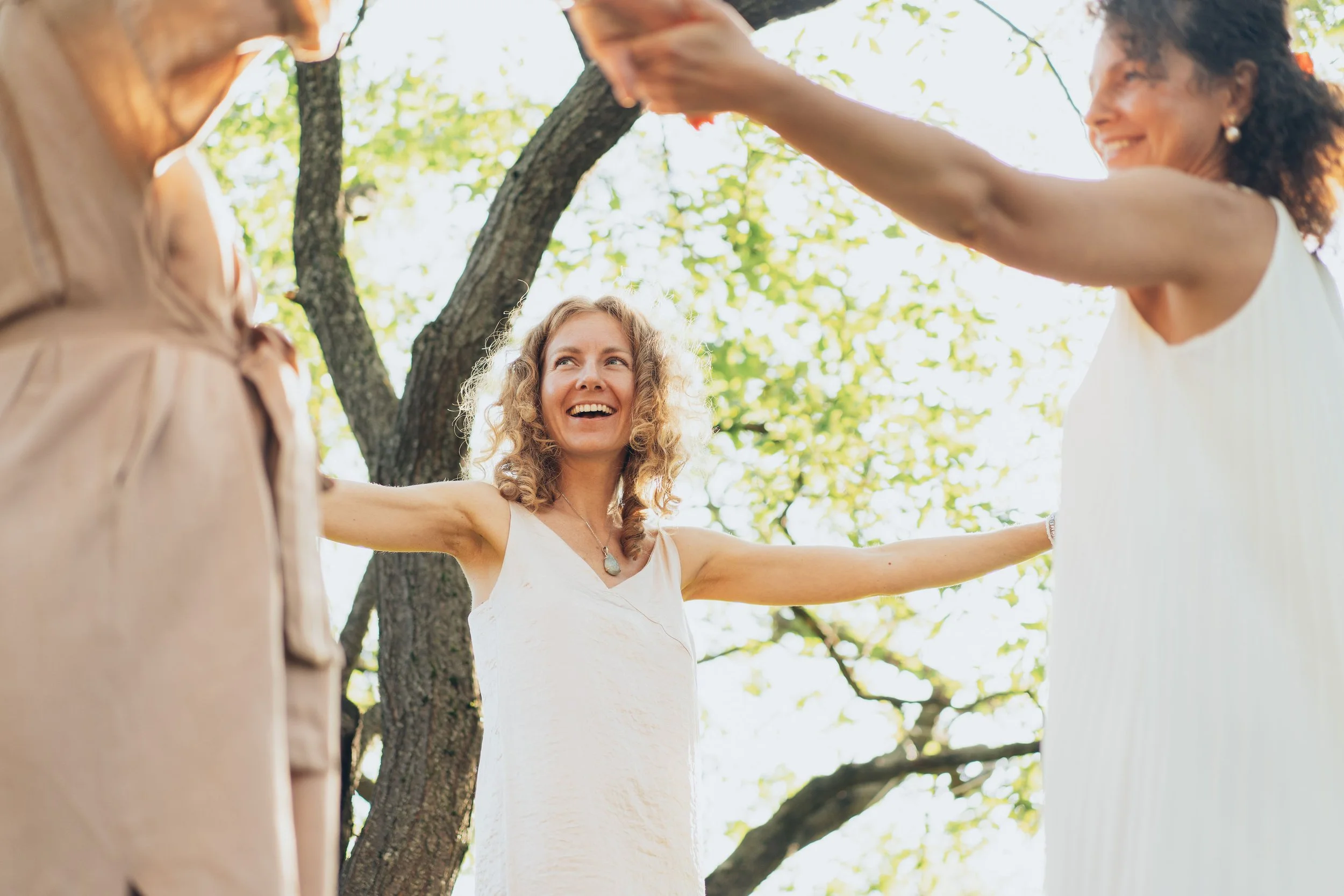 A group of women holding hands in a circle outdoors under a tree on a sunny day, smiling and enjoying each other's company.