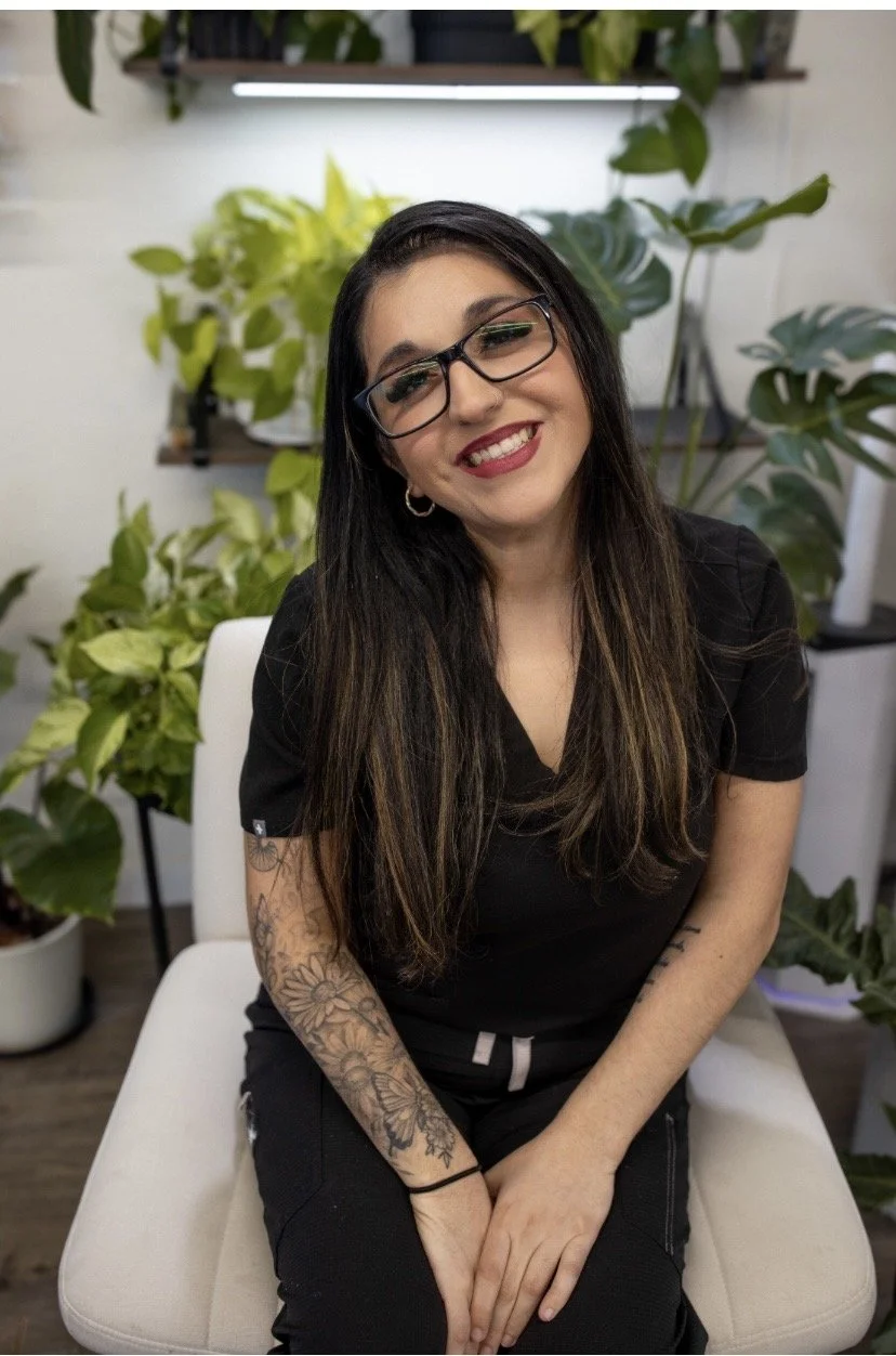 A woman with long dark hair, glasses, and tattoos on her arm, smiling and sitting on a white chair with green plants in the background.