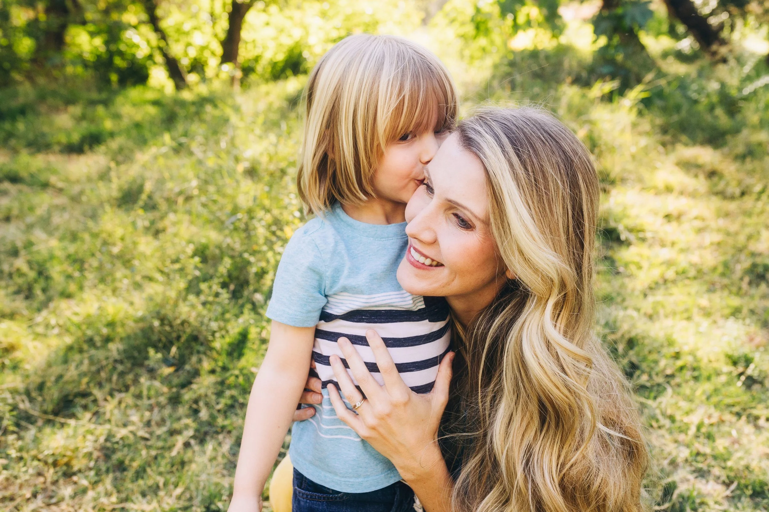 A woman with long blonde hair smiling as she embraces a young boy with straight, blonde hair, outdoors in a forested area with sunlight filtering through trees.