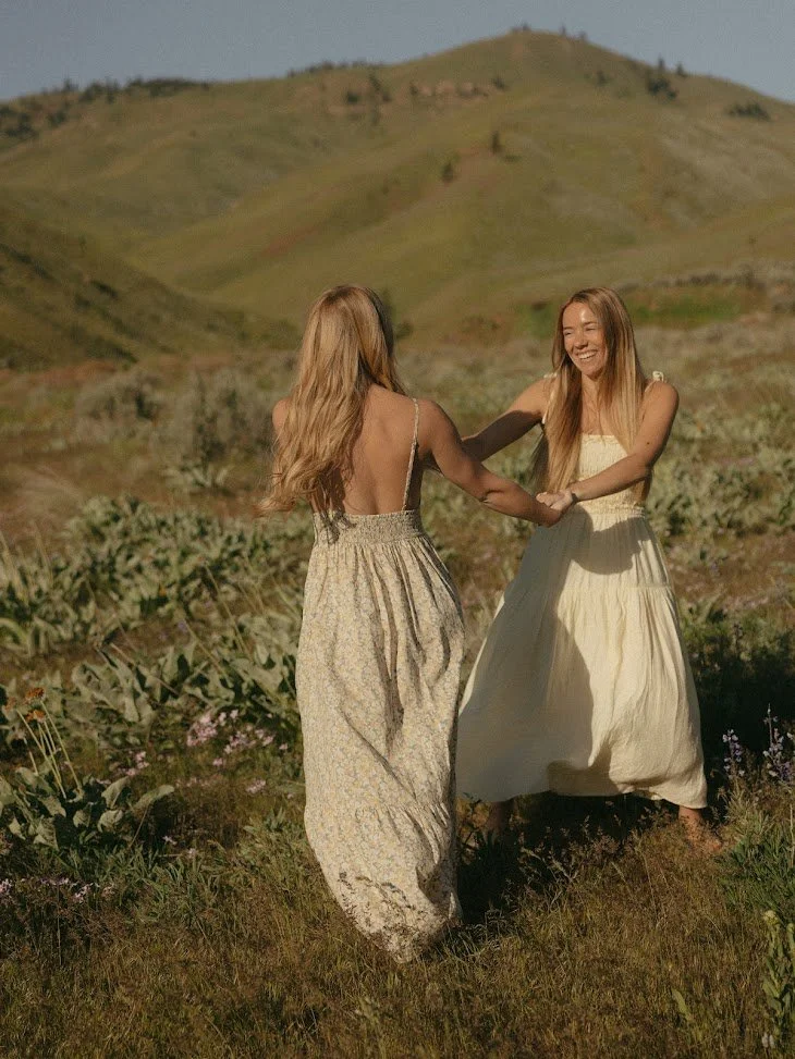 Two women holding hands and laughing in a grassy, hilly outdoor setting during daylight.