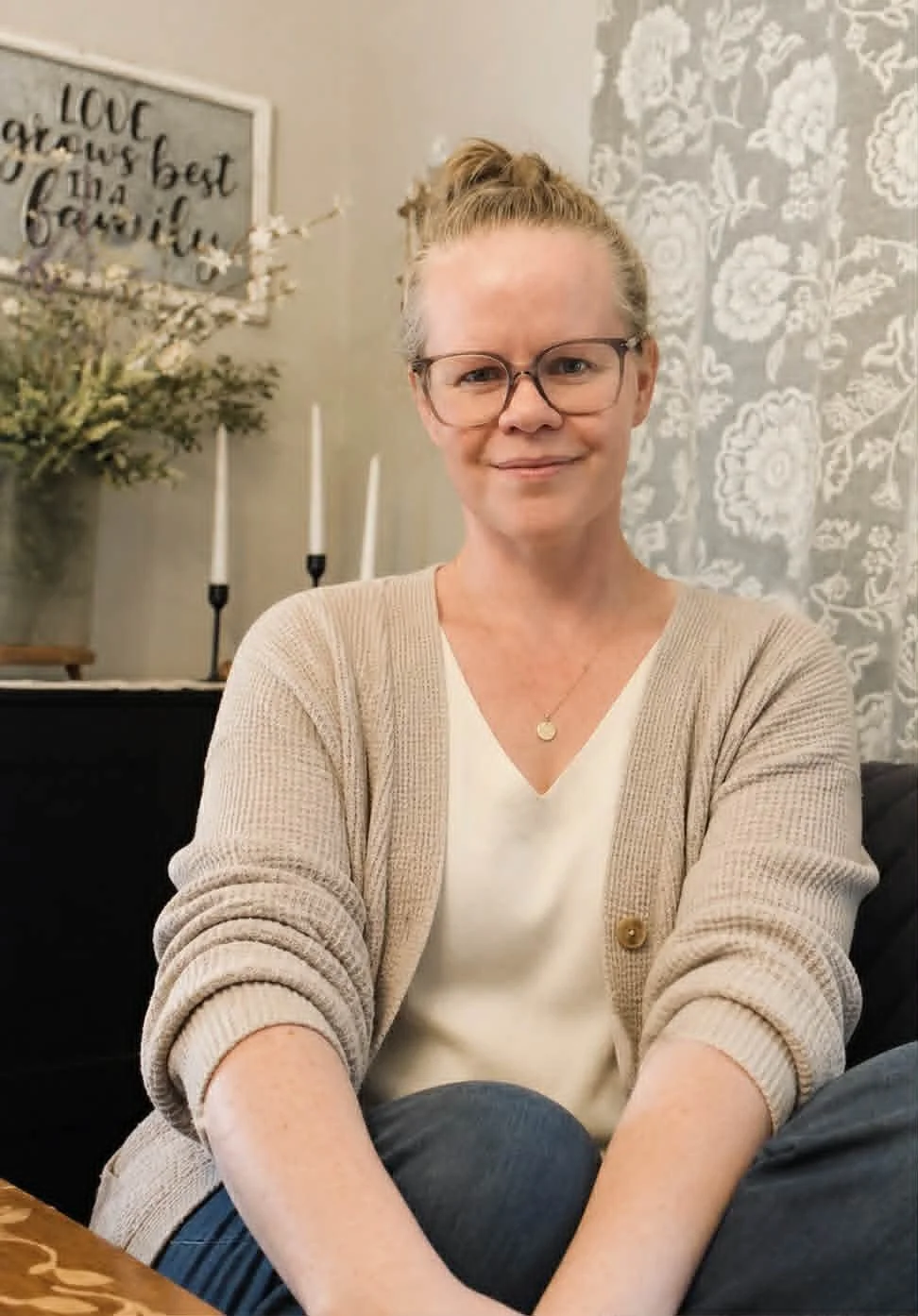 A woman with glasses and a bun hairstyle smiling at the camera, sitting indoors with a beige cardigan, white blouse, and dark pants. In the background, there is a decorative wall, a floral arrangement, and a framed quote.