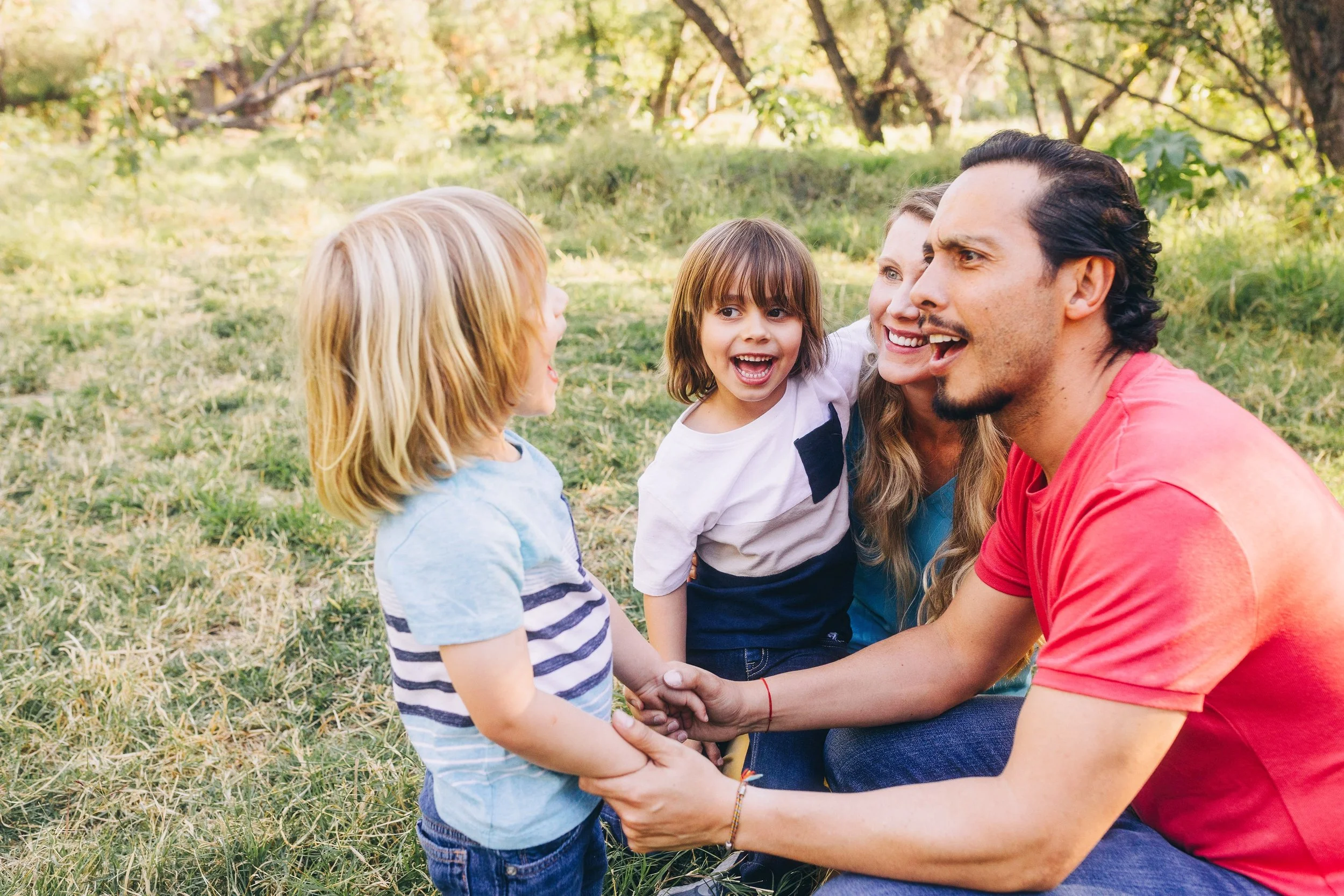 A man and woman kneeling on the grass outdoors, holding hands with a young girl who is smiling, surrounded by trees and greenery.