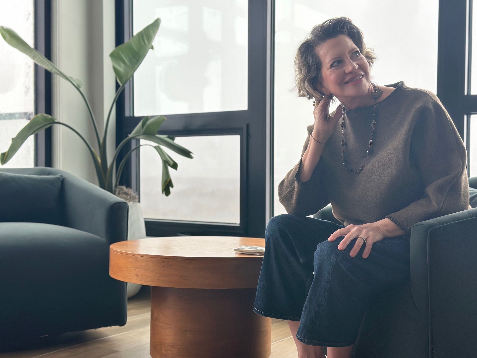 A woman with short, wavy hair smiling and sitting on a chair in a modern, bright room with large windows. There is a green houseplant and a round wooden table nearby.