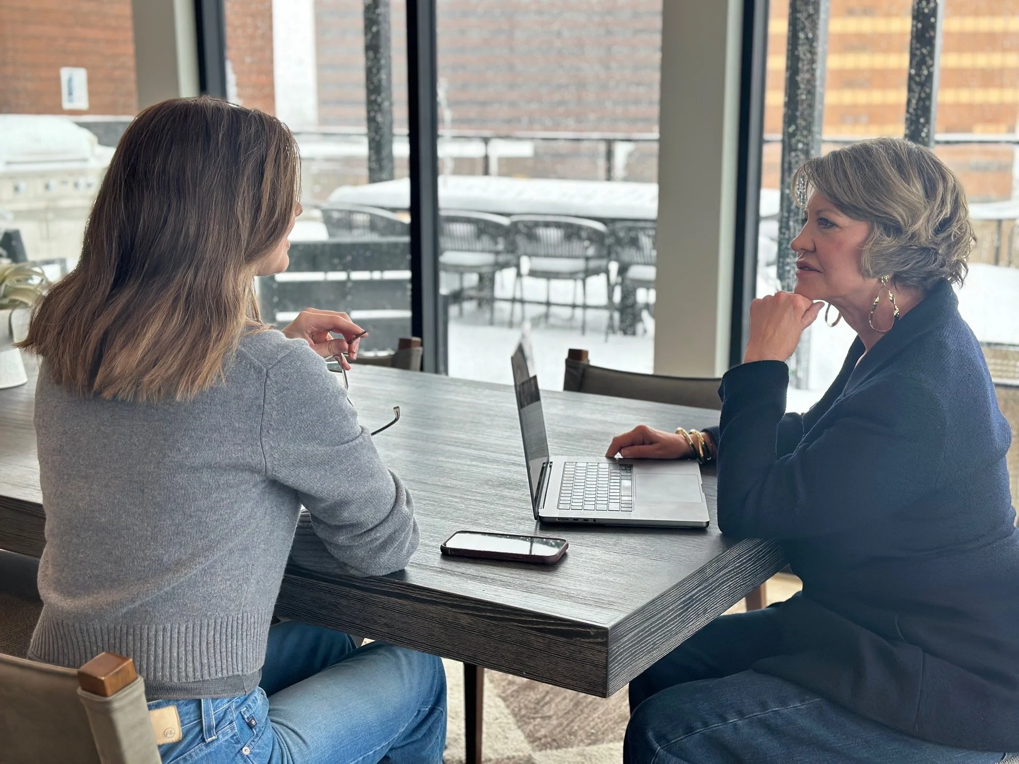 Two women sitting at a table, engaged in conversation. One woman has shoulder-length hair and is wearing a gray sweater, the other has short curly hair and is wearing a black blazer. There is a laptop, a smartphone, and a pair of glasses on the table. The setting is indoors with large windows showing a snowy outdoor scene.