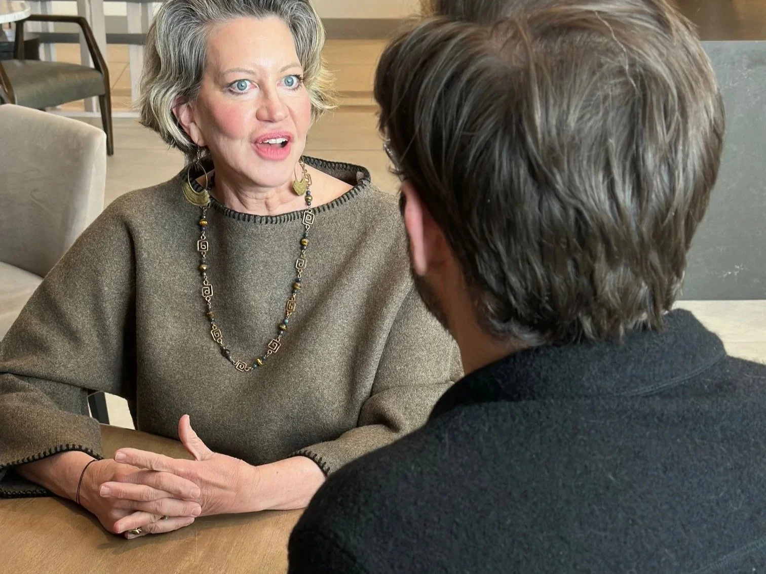 A woman with short gray hair and blue eyes engaged in conversation with a man with brown hair, seen from behind, sitting at a table in a well-lit room.