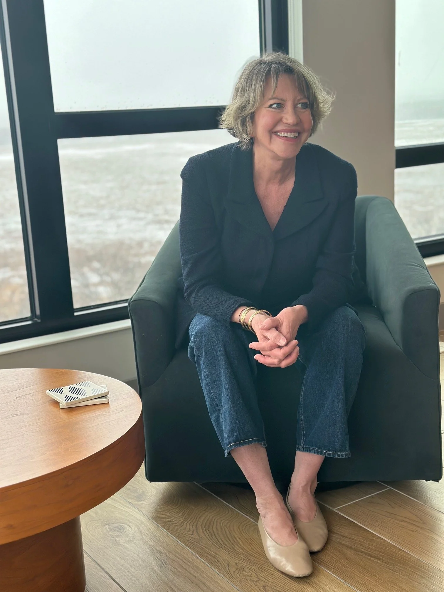 A woman with short blonde hair and a black blazer is sitting barefoot on the floor next to a gray armchair. She is smiling and sitting near a desk with a glass of water and some papers in front of her. There are large windows behind her showing an outdoor scene.