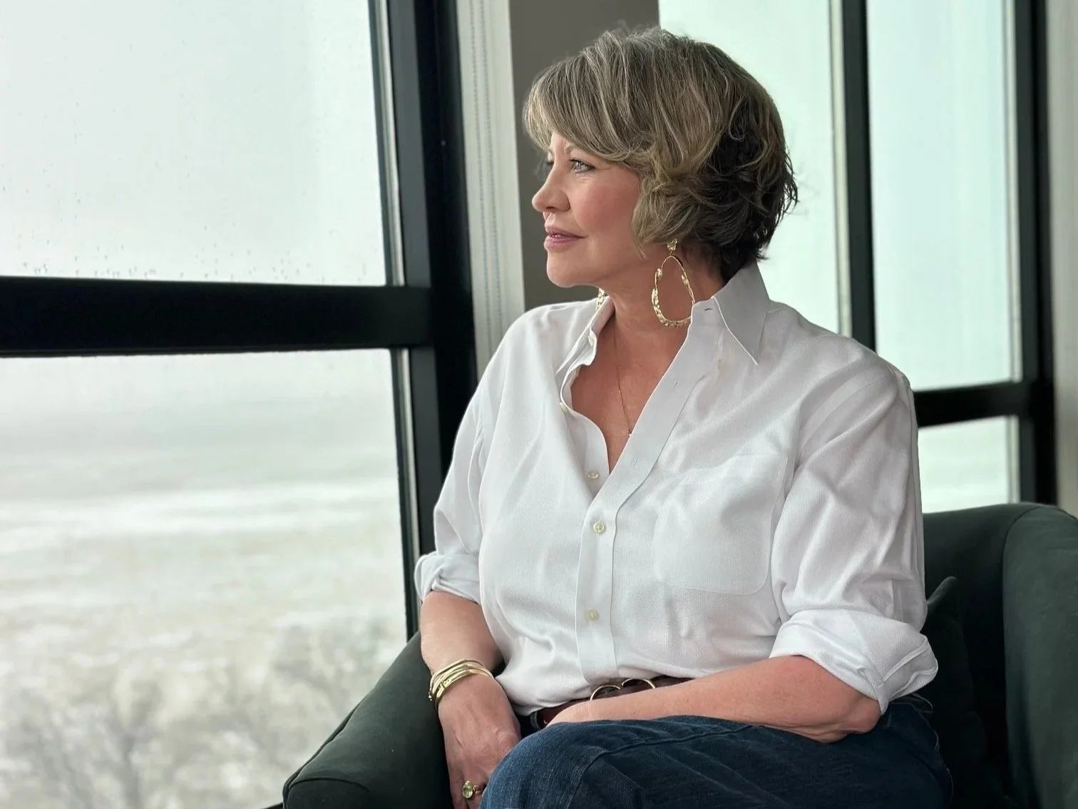 A woman with short, wavy hair wearing a white button-up shirt and gold hoop earrings, seated indoors near large windows overlooking a body of water.