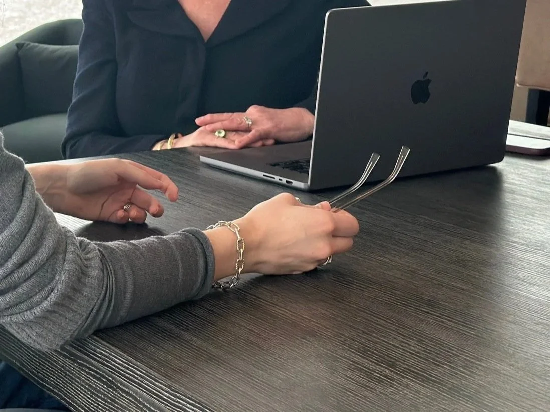 Two people sitting at a dark wooden table, one with a gray sleeve, holding glasses and a bracelet, and the other in a black shirt with bracelets and rings; a silver MacBook laptop on the table.