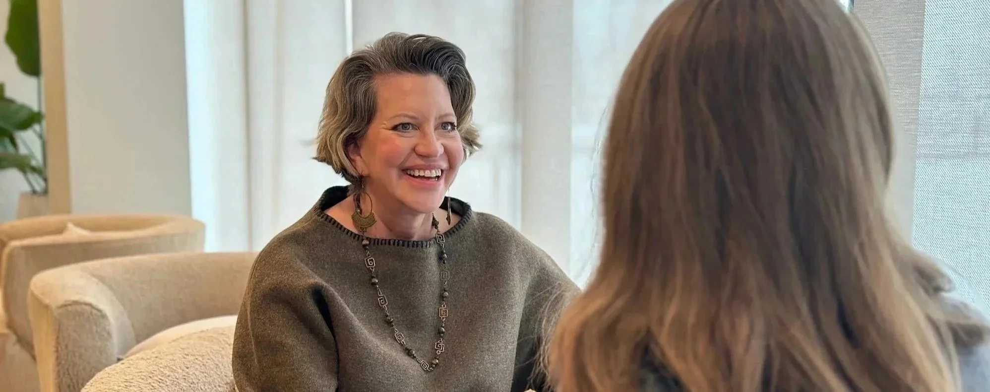 Two women having a conversation in a cozy, well-lit room with beige chairs and a large window. The woman facing the camera has short, wavy hair, colorful earrings, and a sweater, smiling warmly.