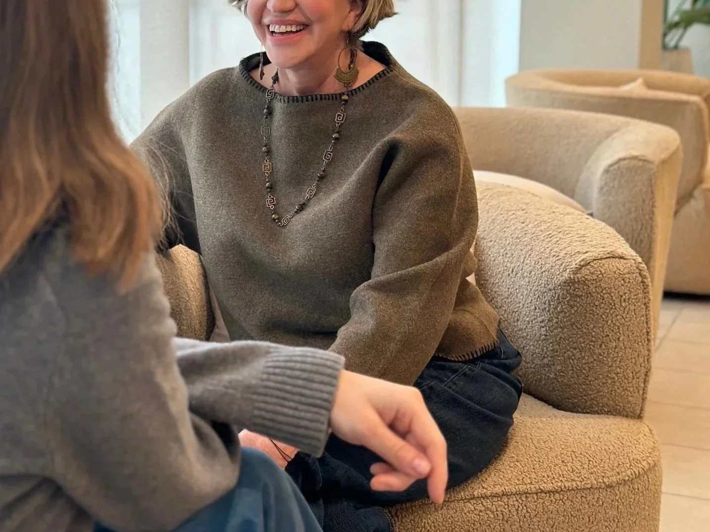 Two women are sitting close together on a beige textured sofa, engaged in a joyful conversation. One woman, with curly hair, is wearing a gray sweater with ribbed cuffs and blue jeans. The other woman, with short blonde hair, is smiling broadly and wearing a brown sweater with a decorative necklace and earrings.