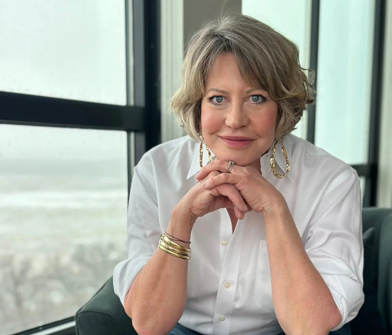 A woman with short, wavy blond hair and blue eyes, wearing a white blouse and large gold earrings, sitting with her hands clasped under her chin, smiling softly at the camera, in a well-lit indoor setting with large windows in the background.