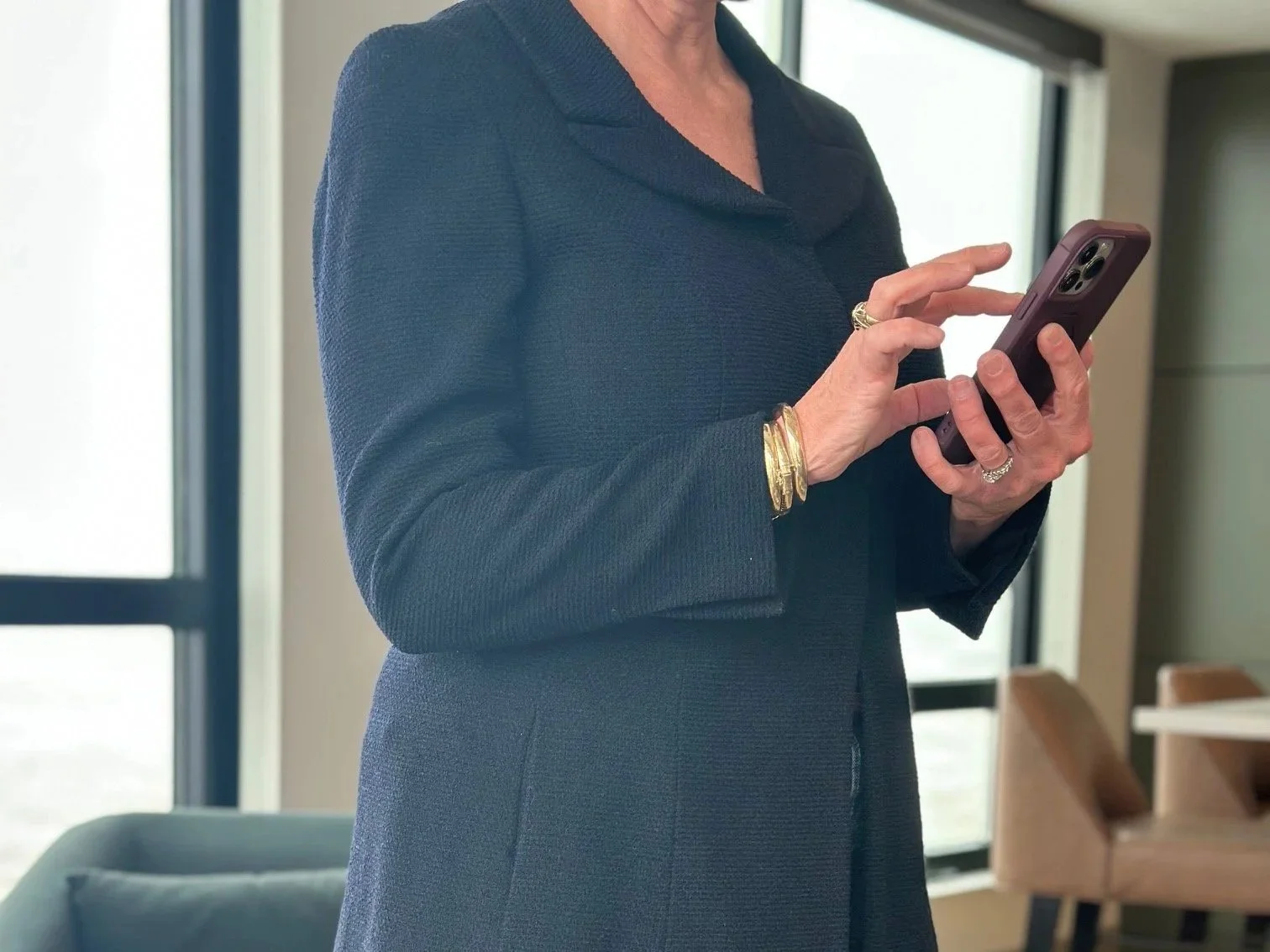 A woman in a dark blazer using a smartphone, wearing multiple gold bracelets and rings, indoors with large windows and modern furniture.