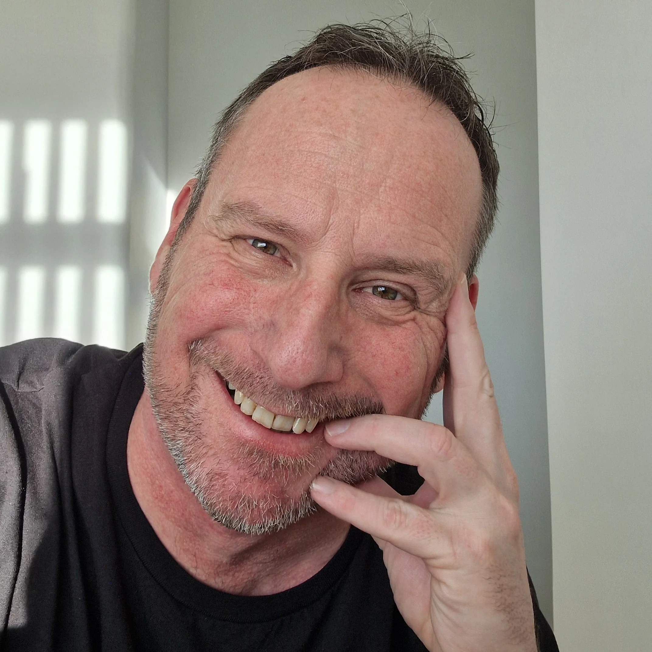 A middle-aged man with a tennis smile, short brown hair, and a trimmed beard, resting his head on his left hand with his fingers touching his cheek, in front of a window with blinds.