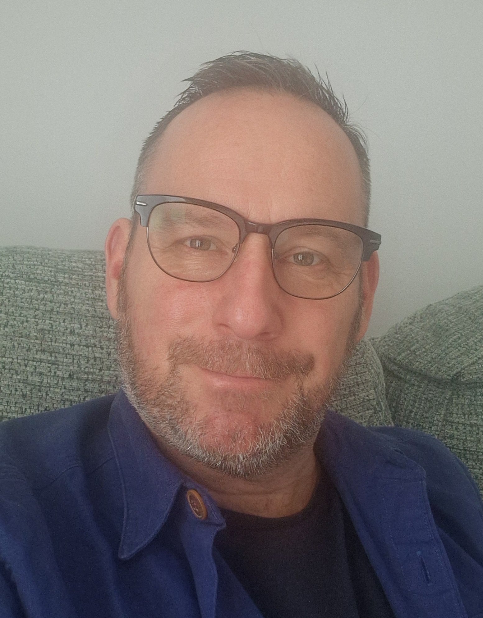 A man with glasses, a beard, and short hair taking a selfie while sitting on a gray couch.