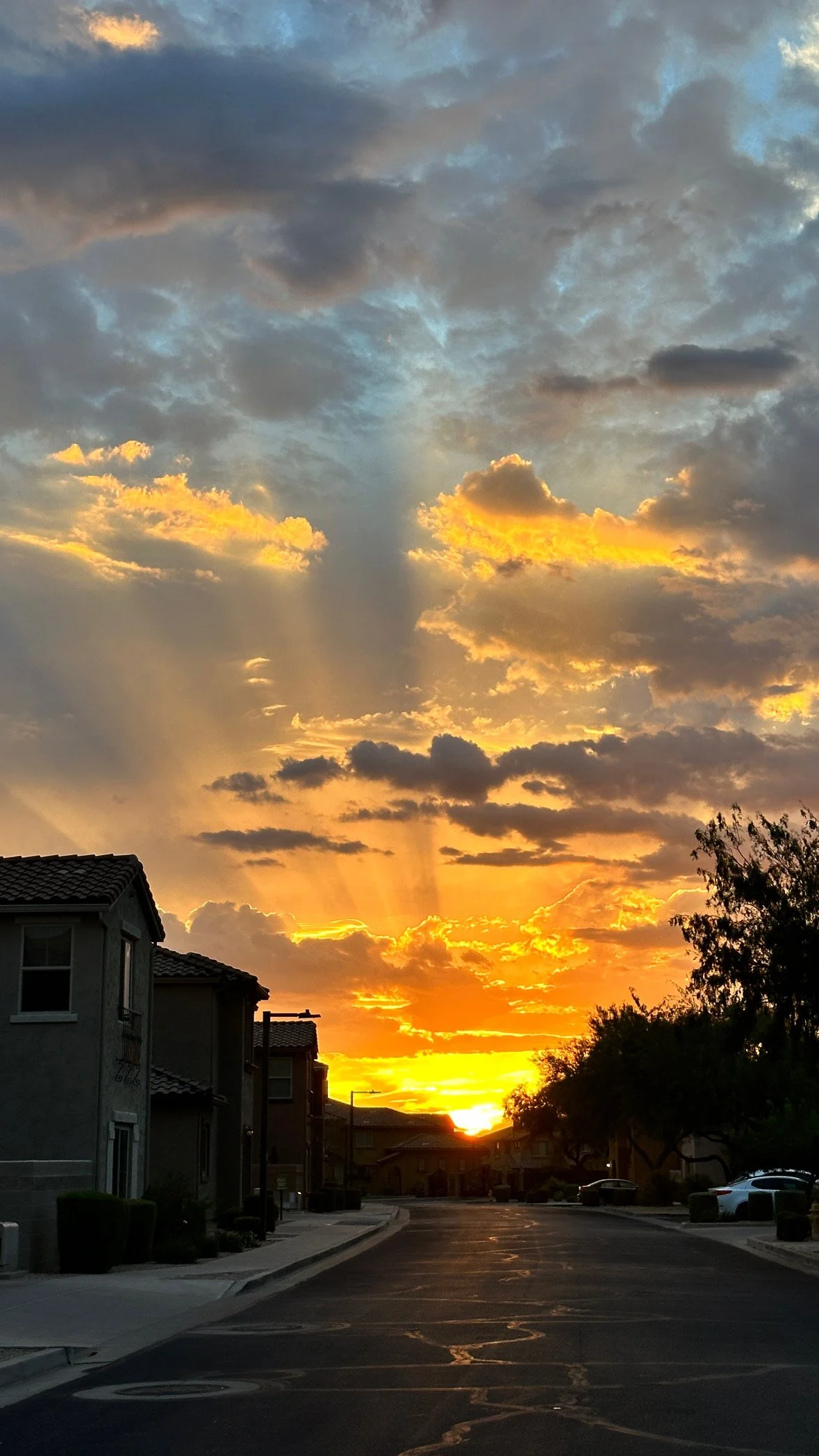 Sunset over a residential street with houses, trees, and parked cars, with clouds and sun rays in the sky.