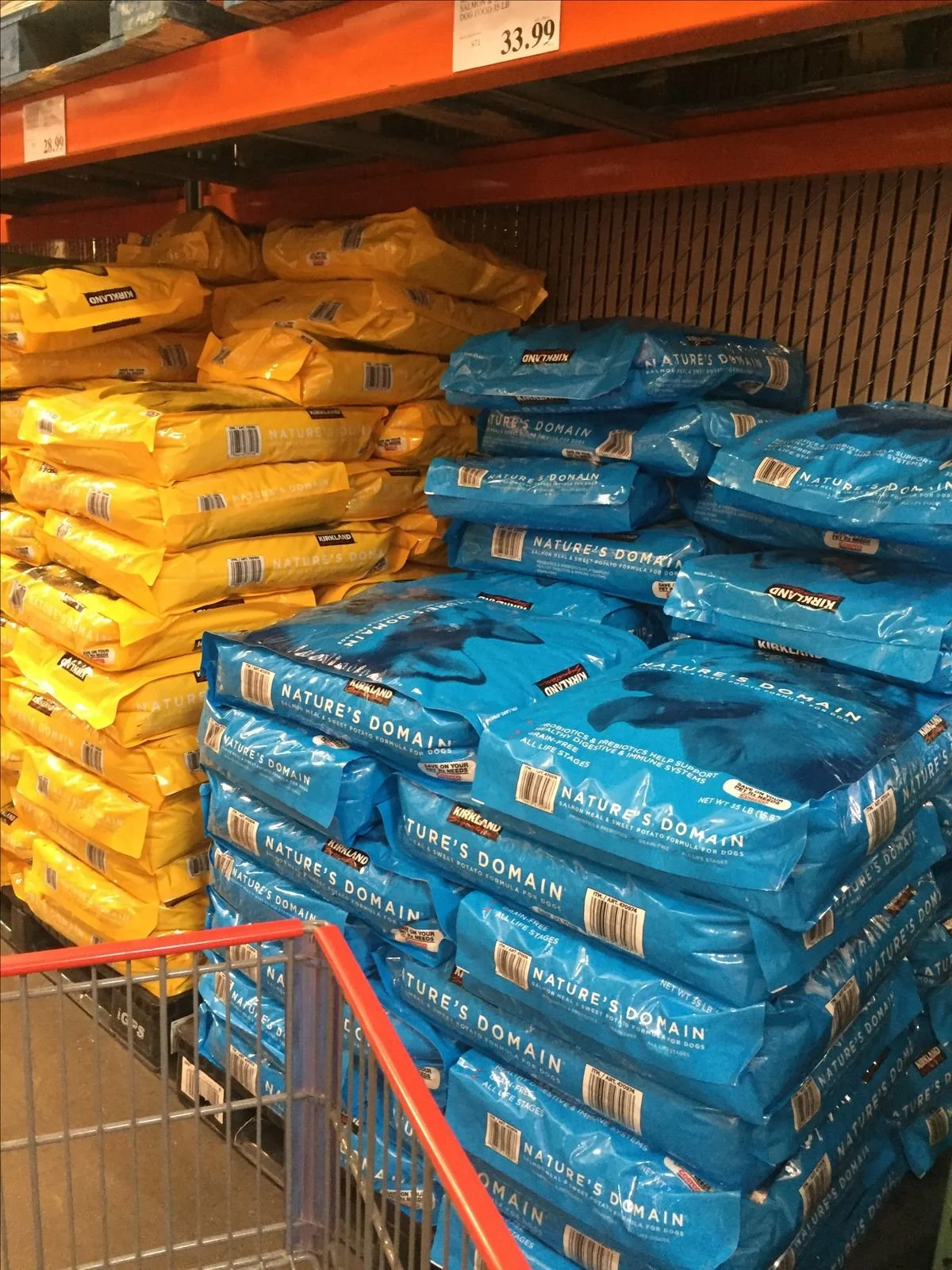 Stacks of yellow and blue bags of dog food on shelves in a store aisle, with a shopping cart in the foreground.