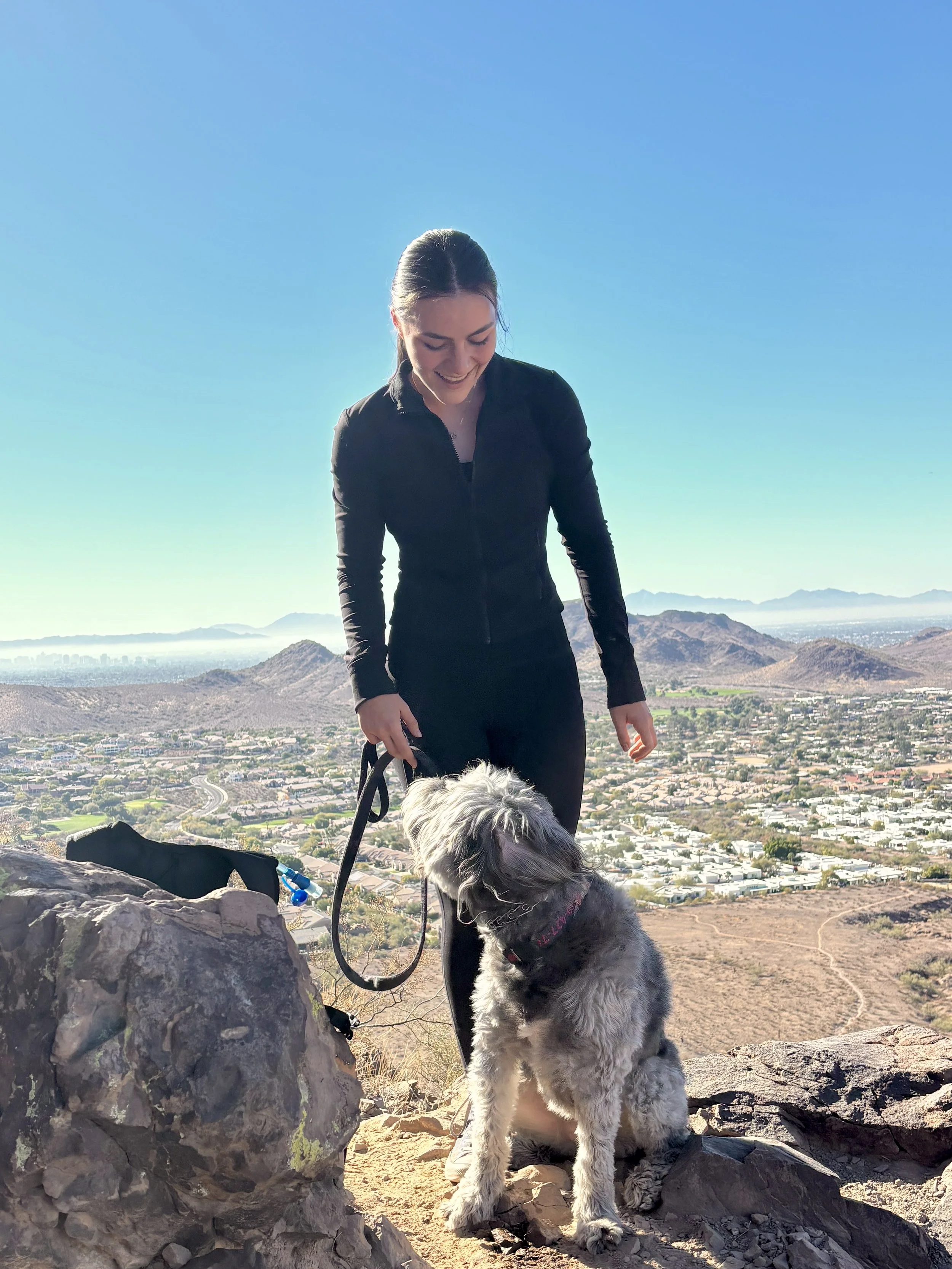 A woman in black athletic clothing with dark hair tied back, smiling while holding a leash attached to a large fluffy gray dog. They are on a rocky hilltop with a sweeping view of a cityscape, mountains in the distance, under a clear blue sky.