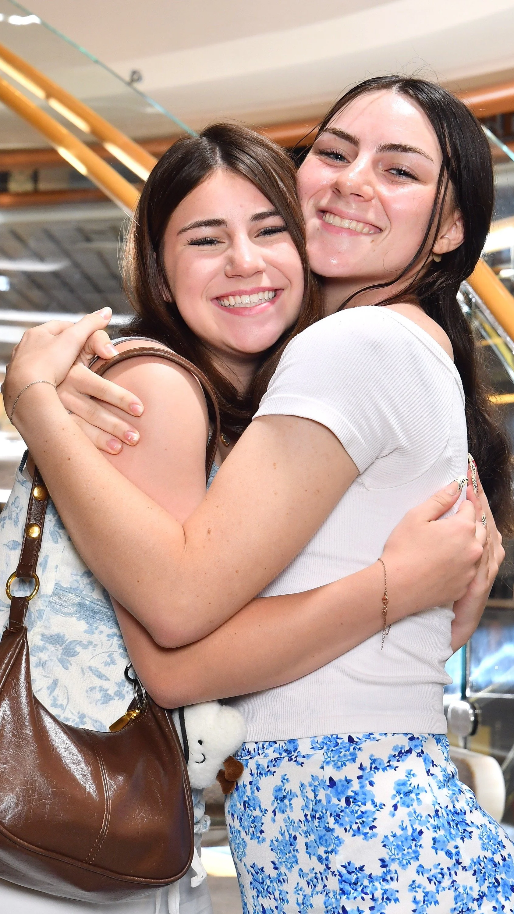 Two young women hugging and smiling indoors, with wooden beams and a staircase in the background.