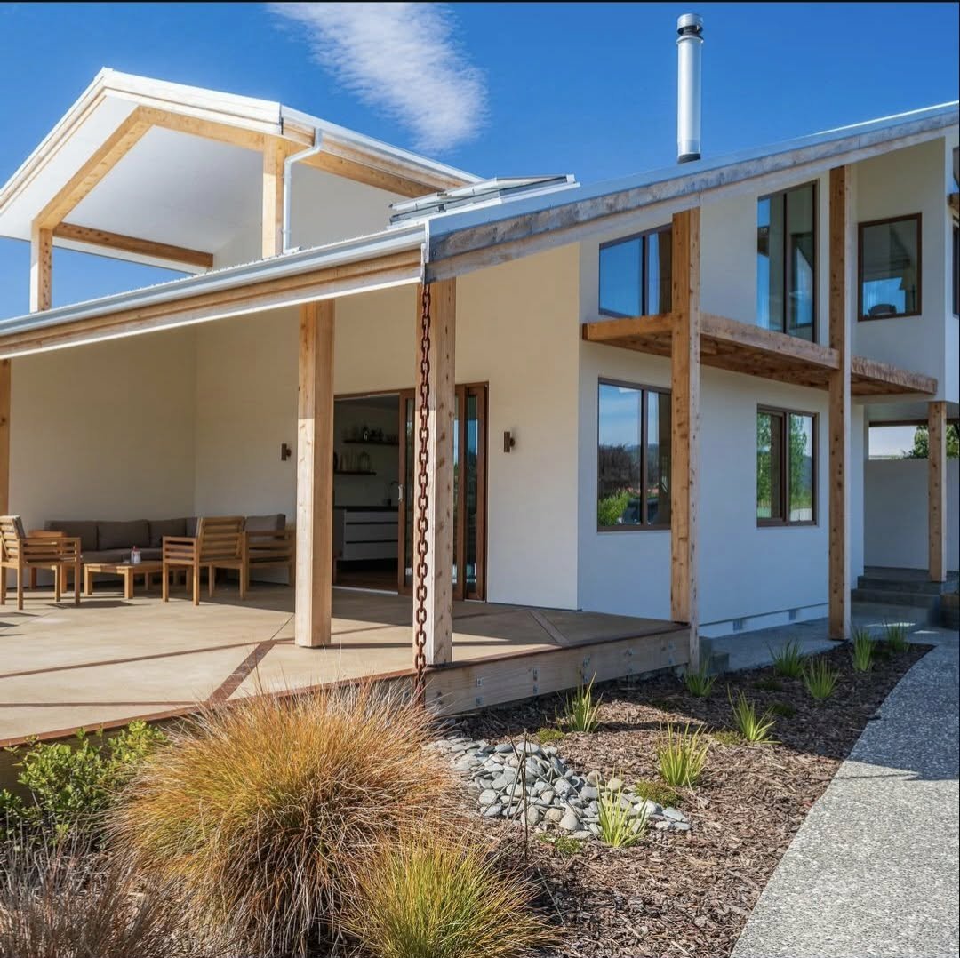Modern two-story house with a wooden deck, outdoor seating, and large windows, situated in a landscaped yard with plants and a concrete pathway.
