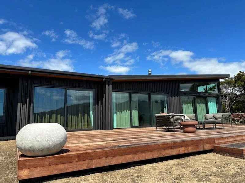 Modern black house with large glass windows and a wooden deck, outdoor seating with wicker chairs and cushions, cloudy blue sky