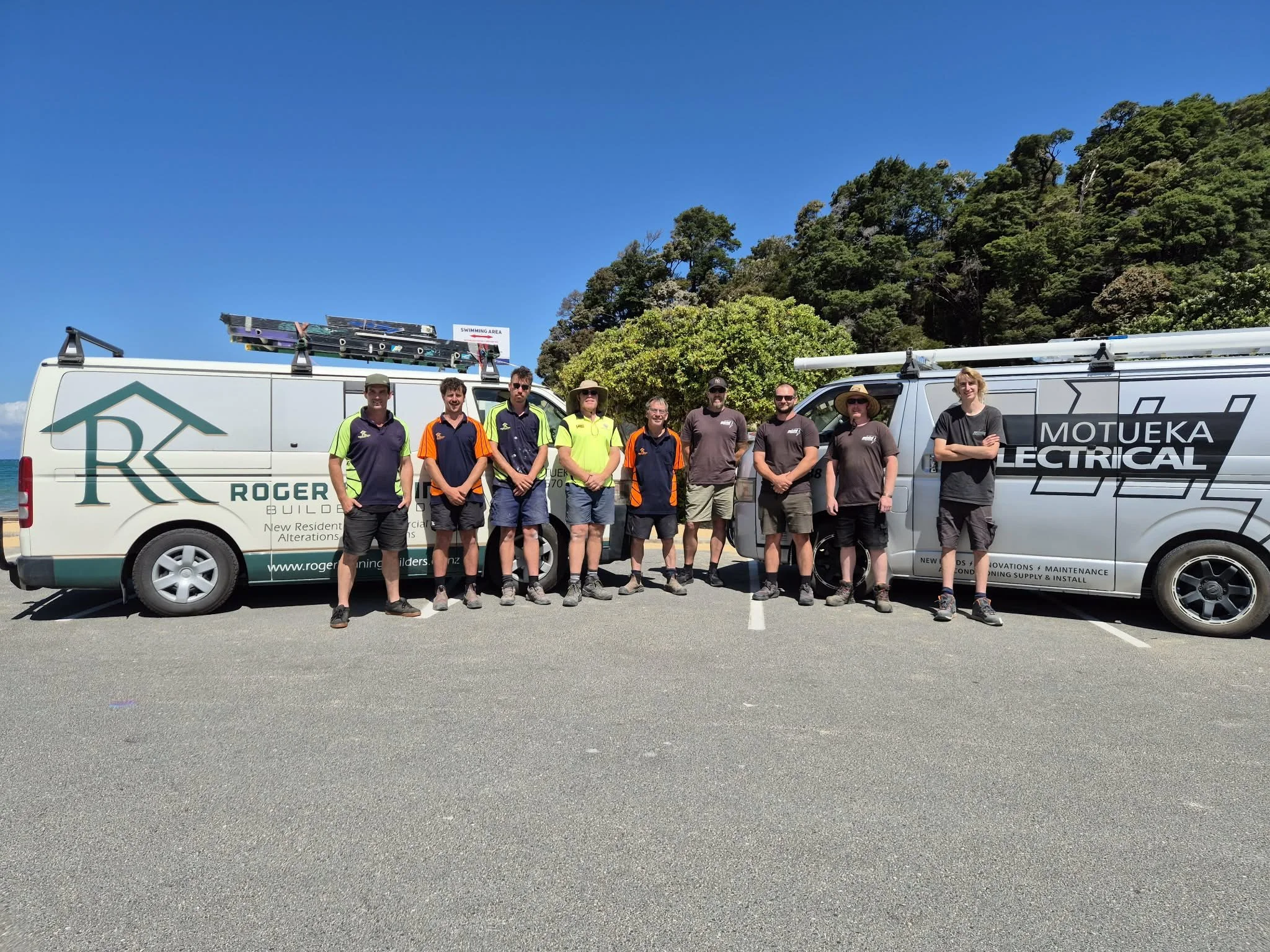 A group of nine people standing in front of two company vehicles with a scenic background of trees and a bright blue sky.