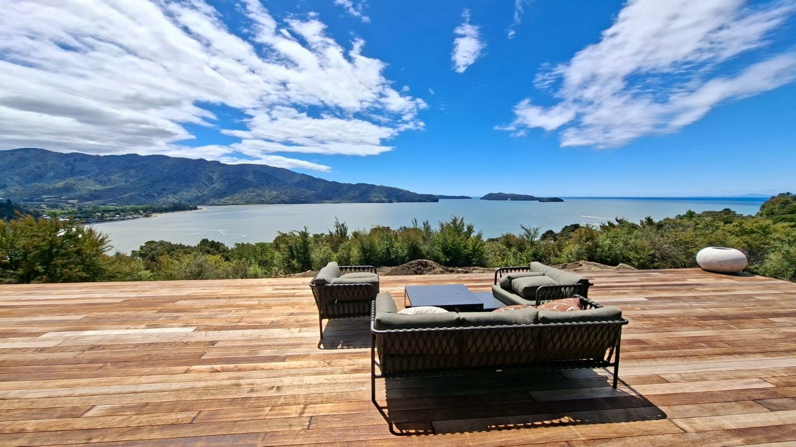 A wooden deck with outdoor furniture overlooking a lake, trees, mountains, and a partly cloudy sky.