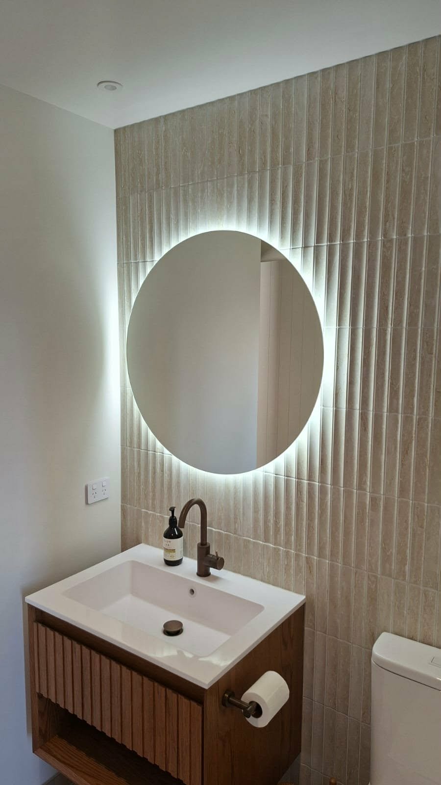 Modern bathroom with a round backlit mirror above a white sink vanity and a bathroom toilet.