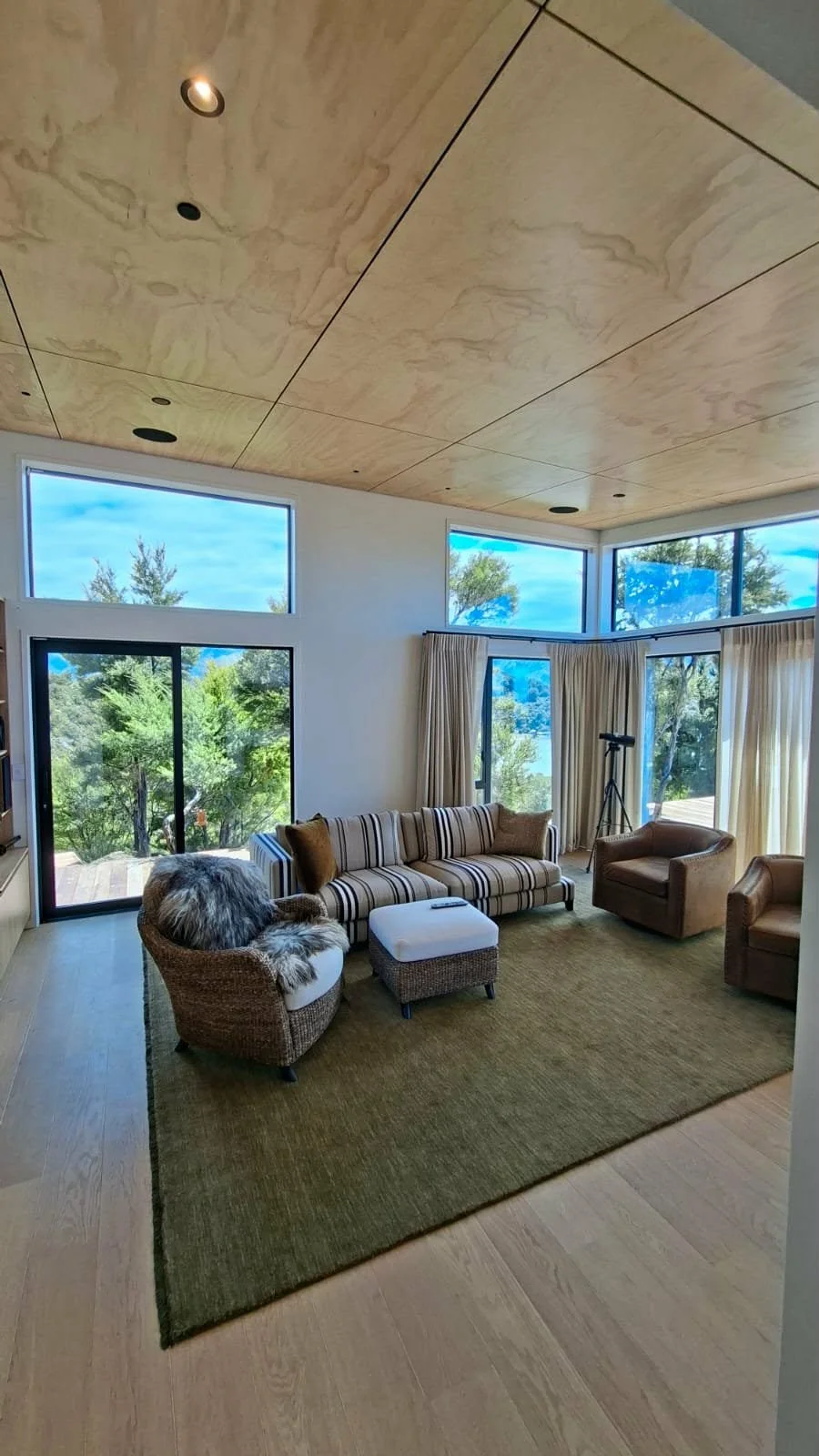 Living room with large windows showing trees outdoors, a beige and brown striped sofa, an armchair with faux fur, another armchair, a ottoman, telescope, and beige curtains on a green rug, with a wooden ceiling.