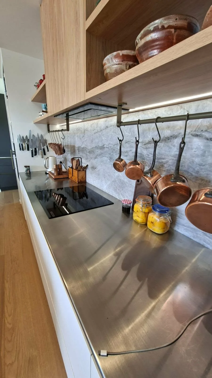 Modern kitchen with stainless steel countertop, copper pots hanging from a rail, wood shelves with ceramic bowls, and a marble backsplash.