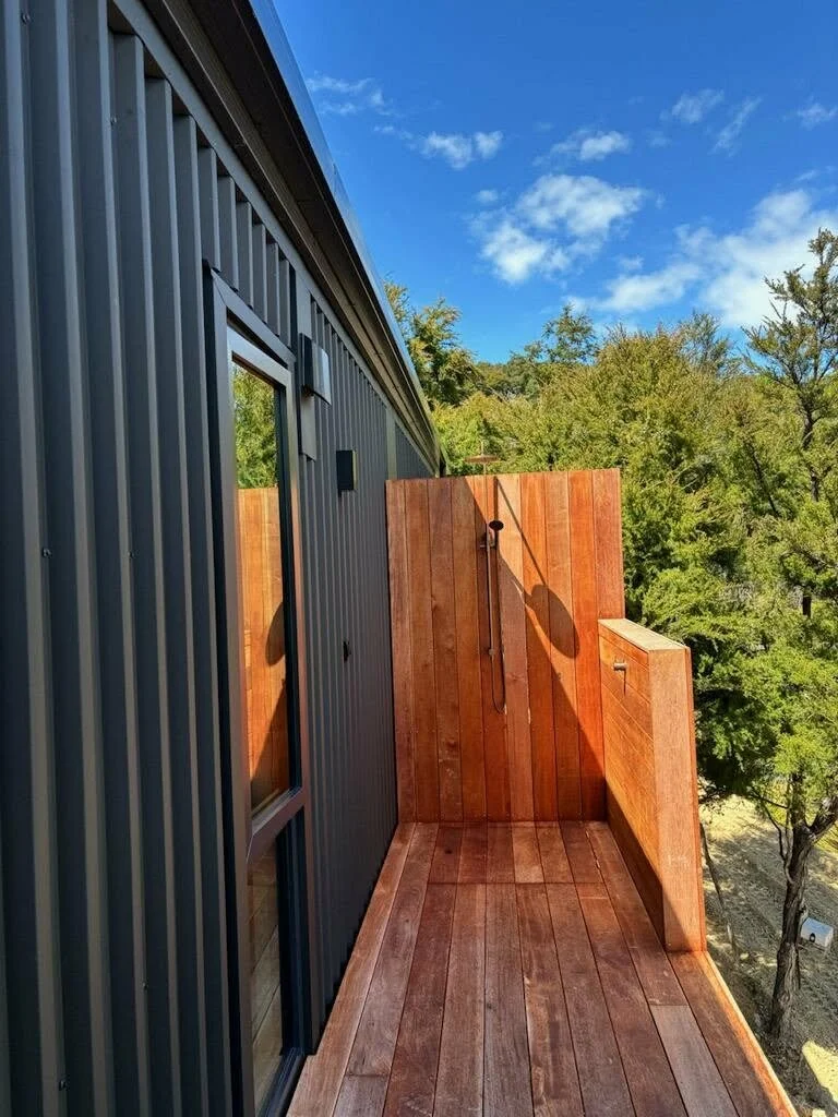 Small outdoor balcony with wooden flooring, privacy wall, and outdoor shower head mounted on the wall, next to a house with metal siding, surrounded by trees under a blue sky with clouds. abel tasman 