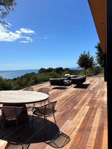 Outdoor patio with a wooden deck, round table with chairs, and lounge chairs, overlooking a body of water under a blue sky with clouds. split apple rock Abel tasman