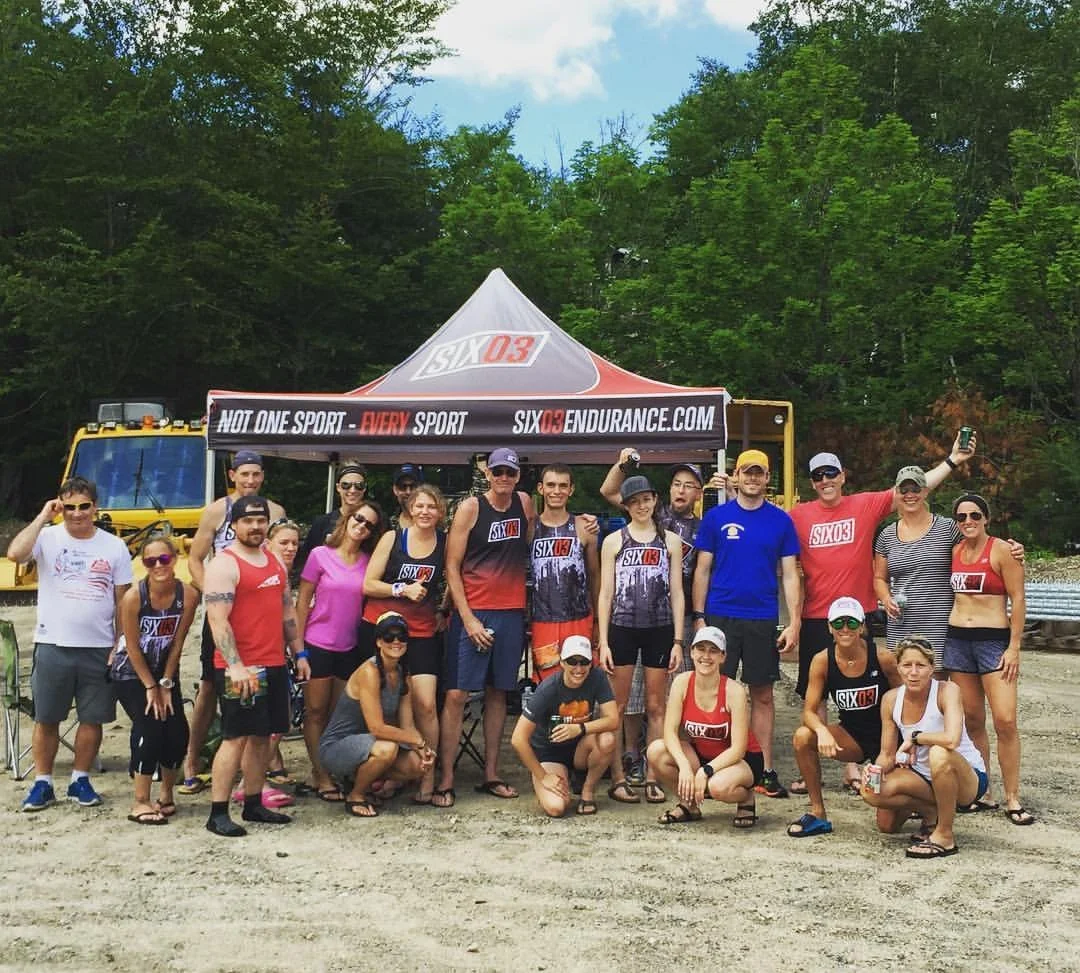 Group of people posing outdoors near a yellow vehicle and a black tent with 'Six03 Endurance' branding. They are wearing athletic and casual clothing, some with matching team shirts, and are smiling at the camera.