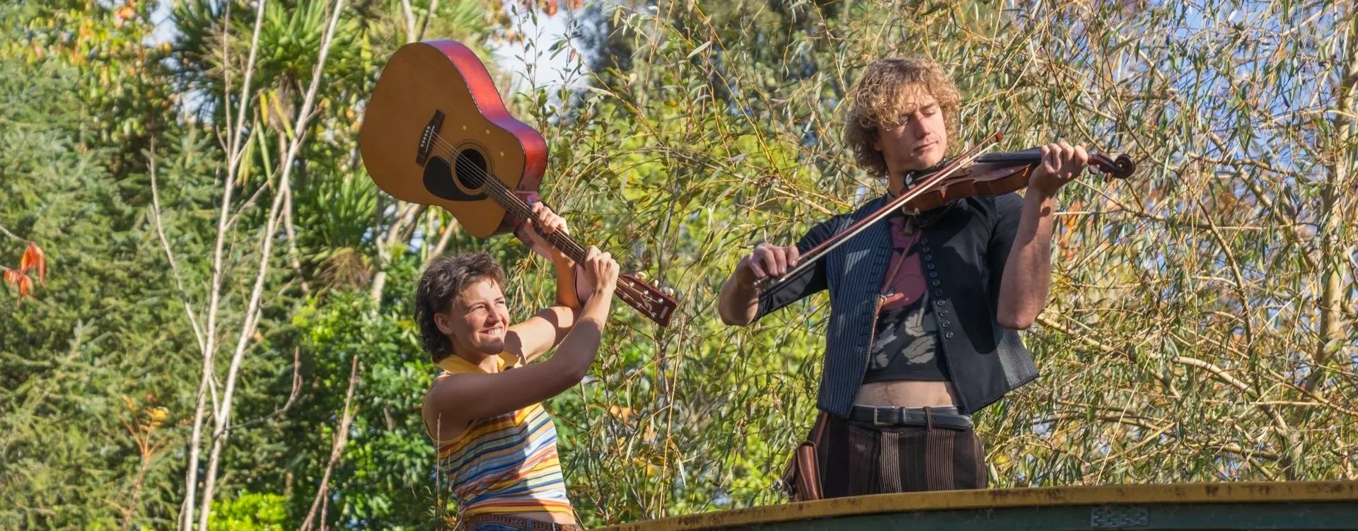 Two people playing musical instruments outdoors among trees. One person is holding a guitar, and the other is playing a violin.