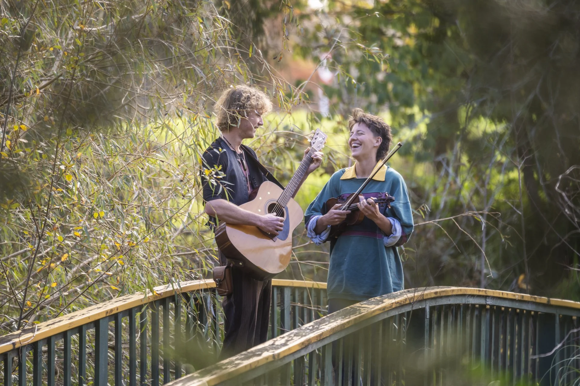 Two women are standing on a wooden bridge outdoors, playing musical instruments and smiling at each other. One woman is playing a guitar, and the other is playing a violin.