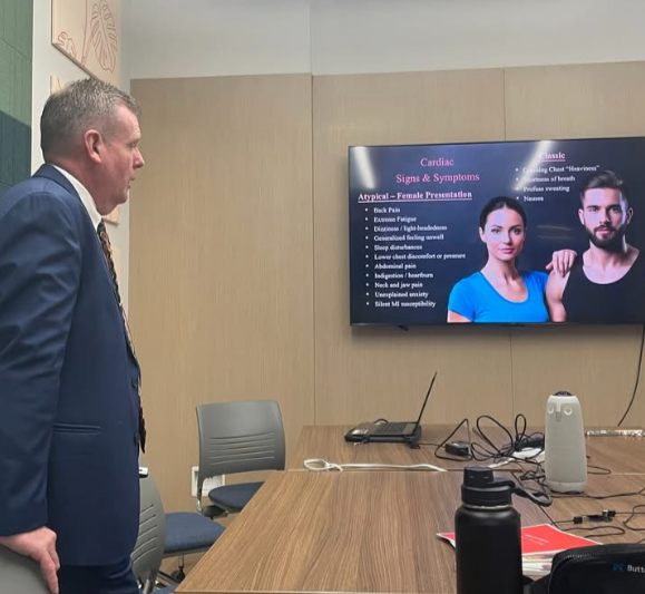 A man in a suit is standing in a conference room, looking at a screen displaying a presentation on cardiac symptoms and signs.