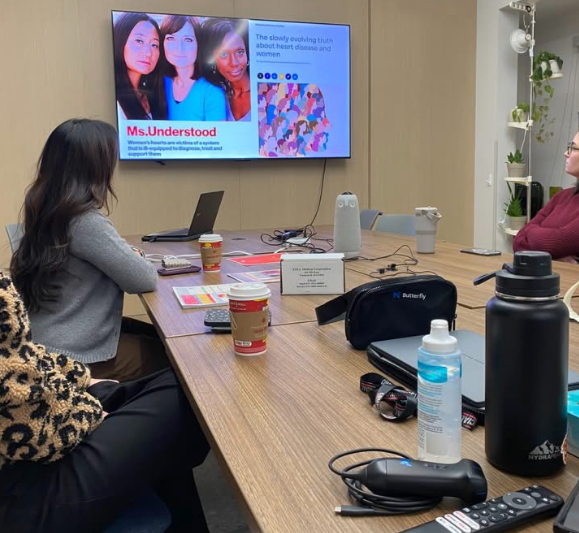 A group of women sitting around a conference table watching a presentation on a large screen. The screen displays images of women and text about women's health. The table has coffee cups, a laptop, and various office supplies.