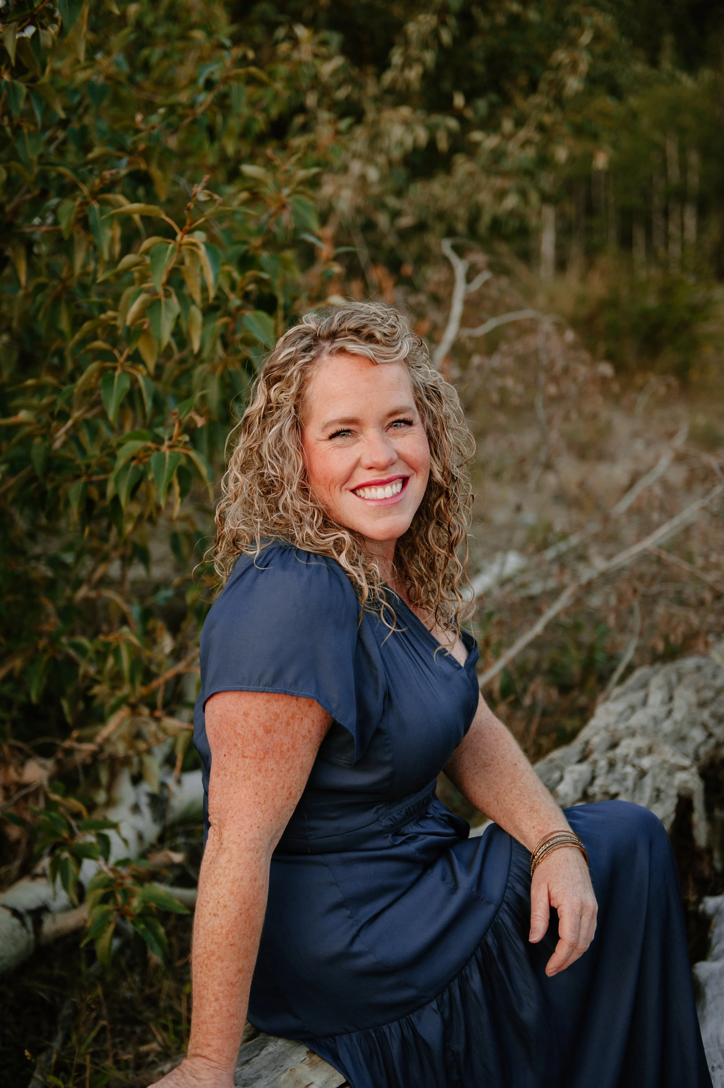 A woman with curly blonde hair, wearing a navy dress, sitting outdoors on a log, smiling at the camera with a background of trees and green foliage.
