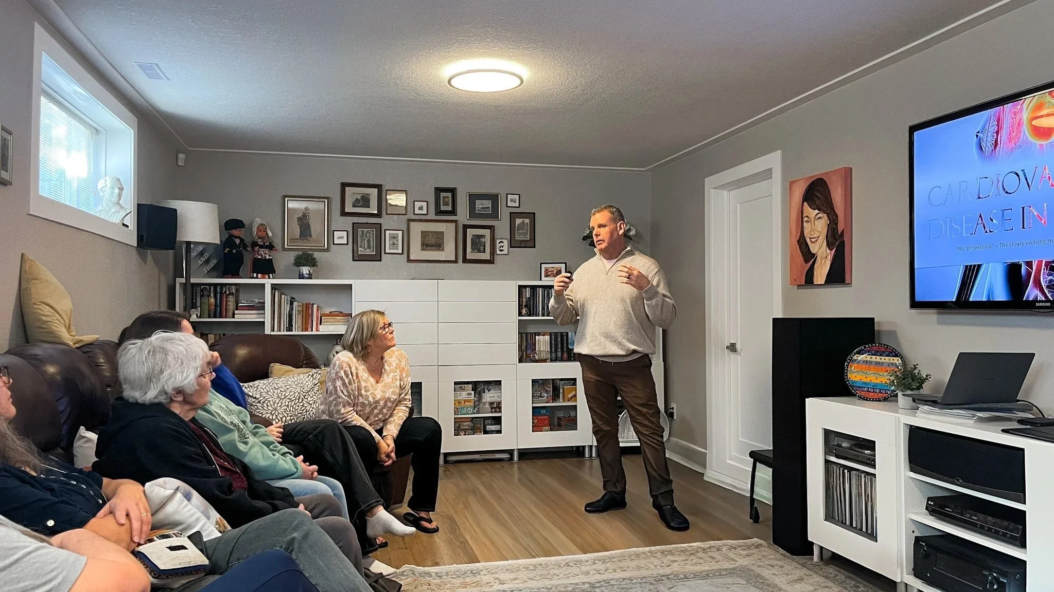 A man giving a presentation to a group of people in a living room setting, with a large screen displaying a medical presentation about heart disease.