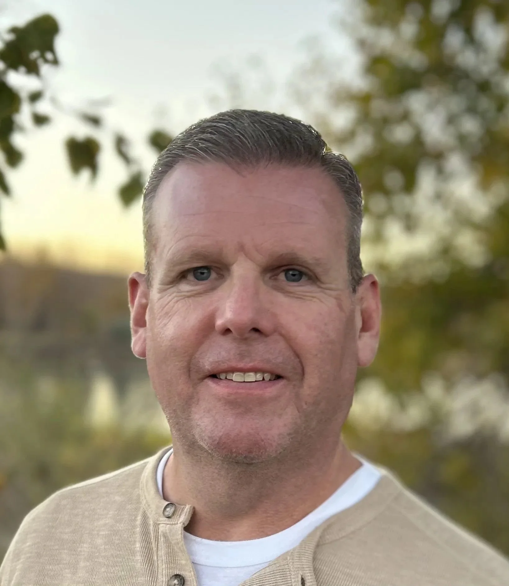 Jim with short dark hair, fair skin, and blue eyes smiling outdoors, with trees and blurred background.