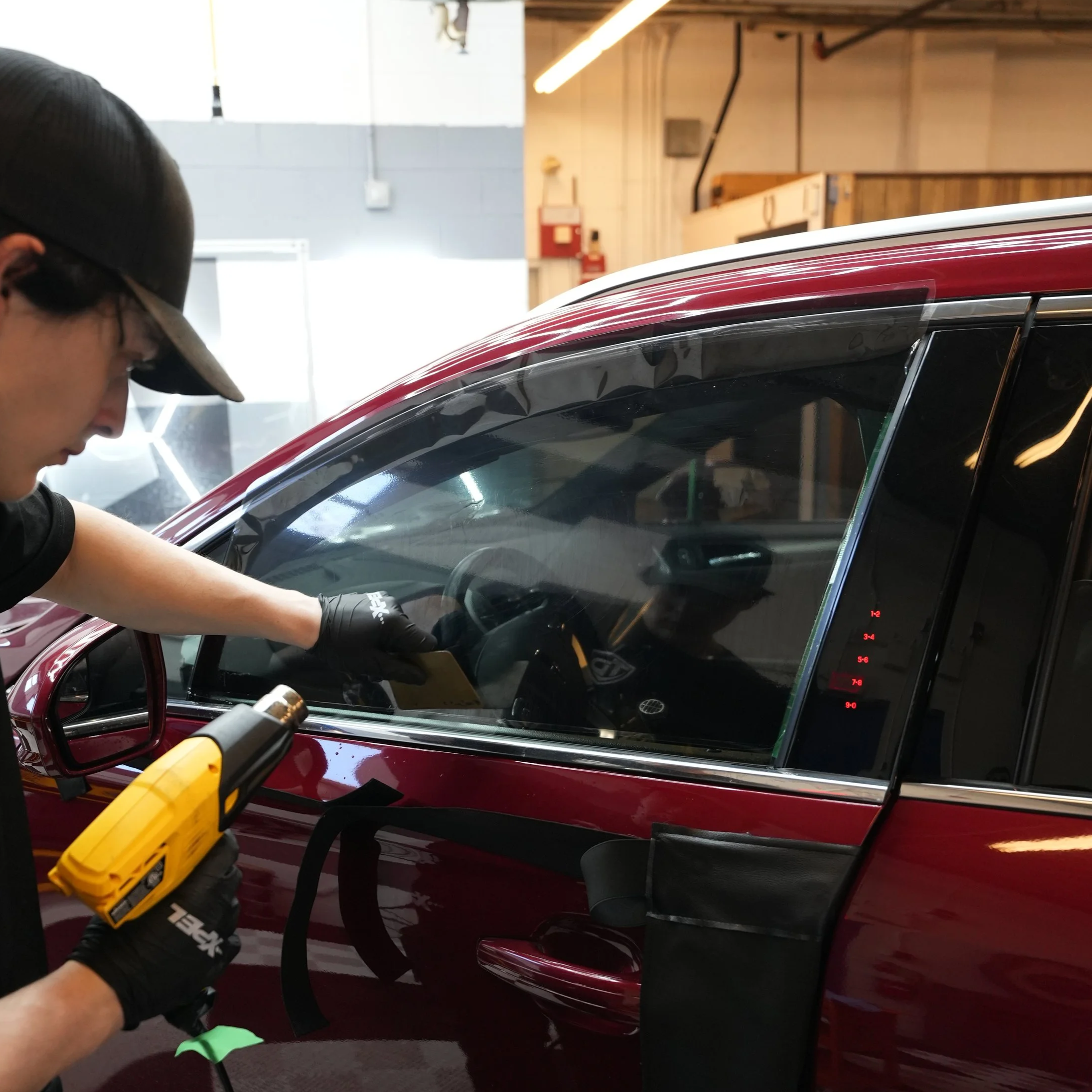 A person wearing black gloves and a baseball cap using a heat gun to work on the exterior of a shiny red car in an indoor workshop.