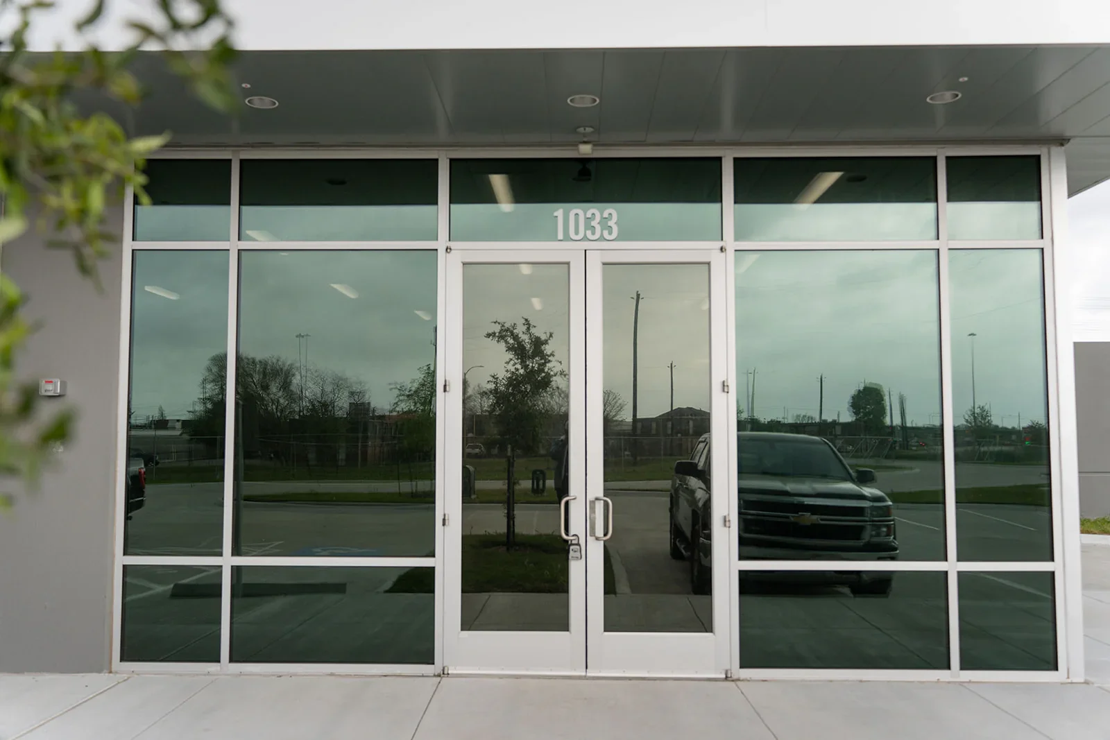 Reflective glass storefront with the number 1033 above the doors, some cars and trees reflected in the glass, and a sidewalk in front.