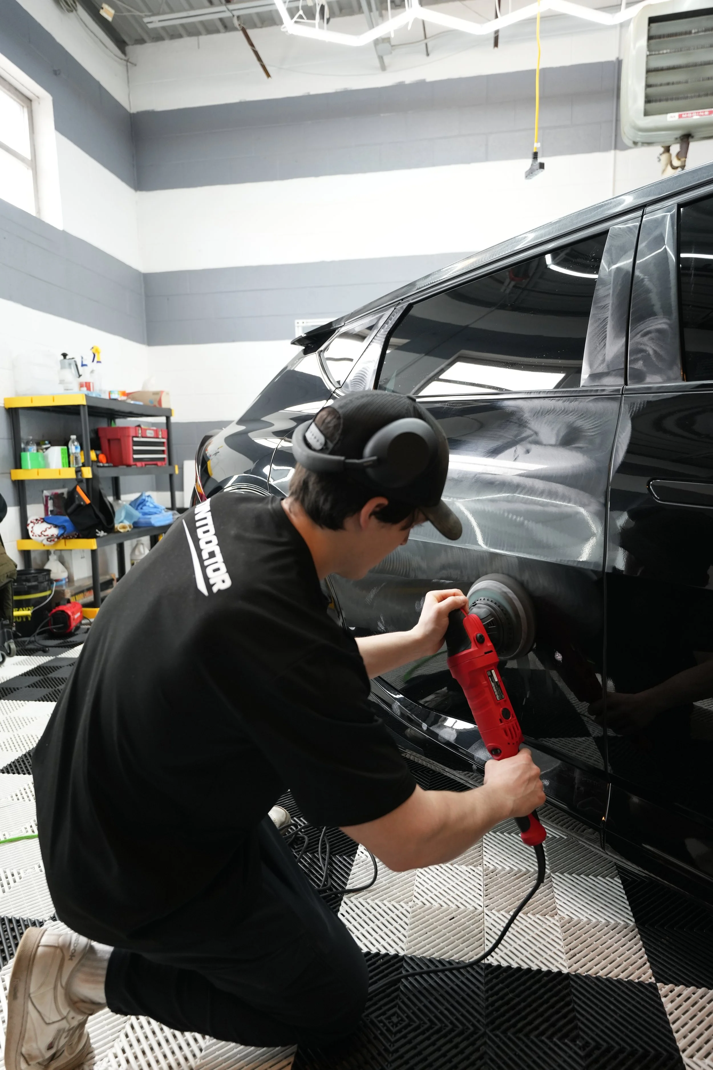 Person kneeling and polishing a black car with a rotary buffer in a garage.