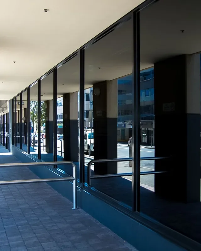 Empty sidewalk with large glass windows reflecting parked cars and buildings, with metal railing in foreground and concrete columns supporting the building overhead