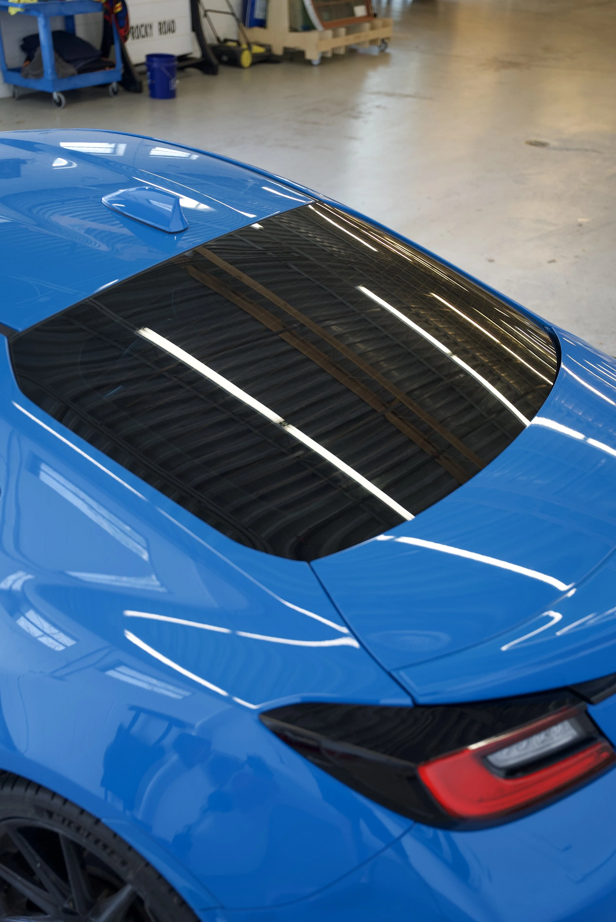 Close-up of a blue sports car with a sleek roof and tinted rear window, parked inside an industrial warehouse or garage.