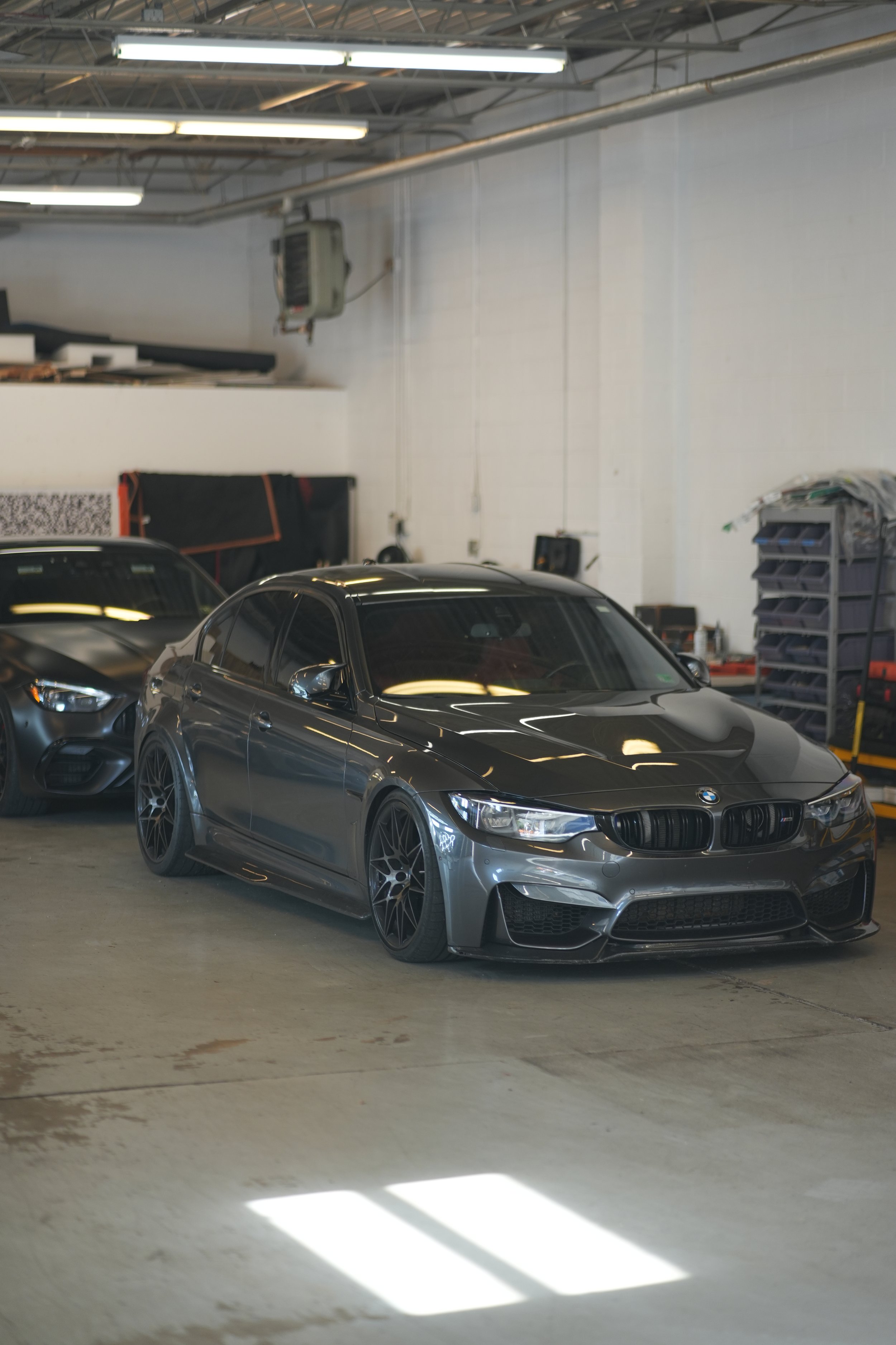 Gray BMW sports car parked in an indoor garage. Other vehicles and storage shelves are visible in the background.