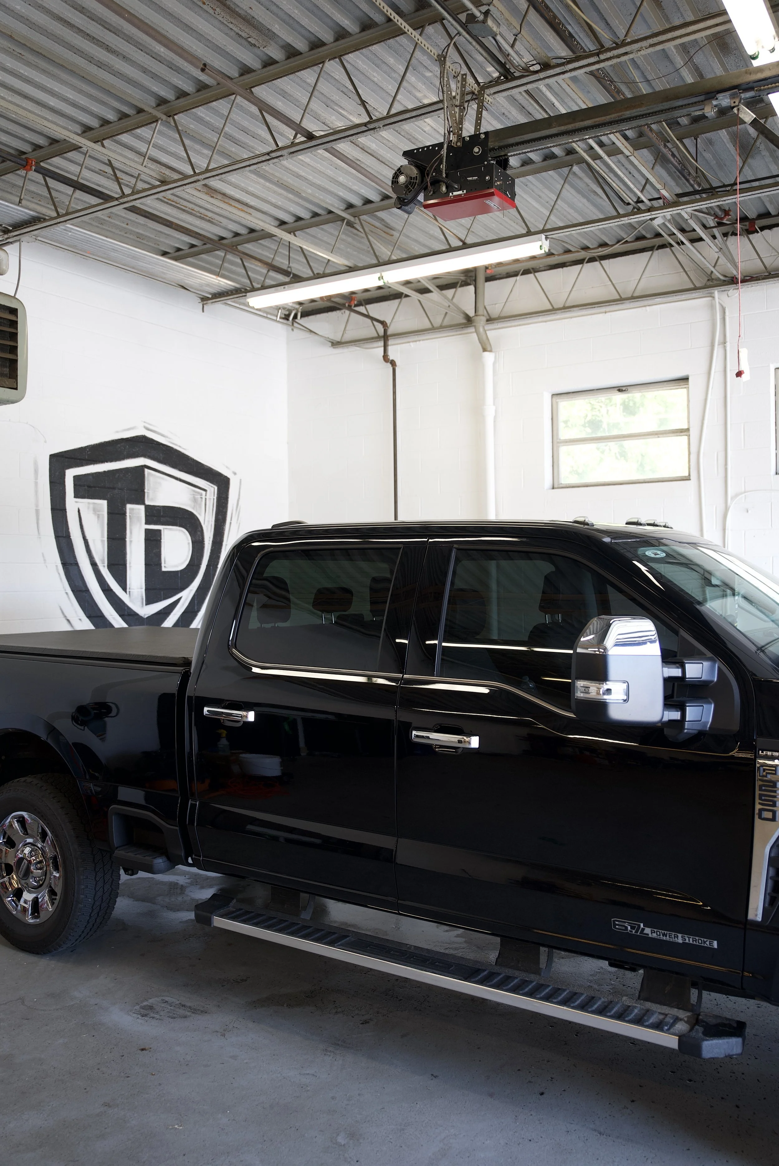 A black pickup truck parked inside a garage with white walls, a metal roof, and a window. Part of a large logo with a shield shape and the letters 'TP' is painted on the wall behind the truck.