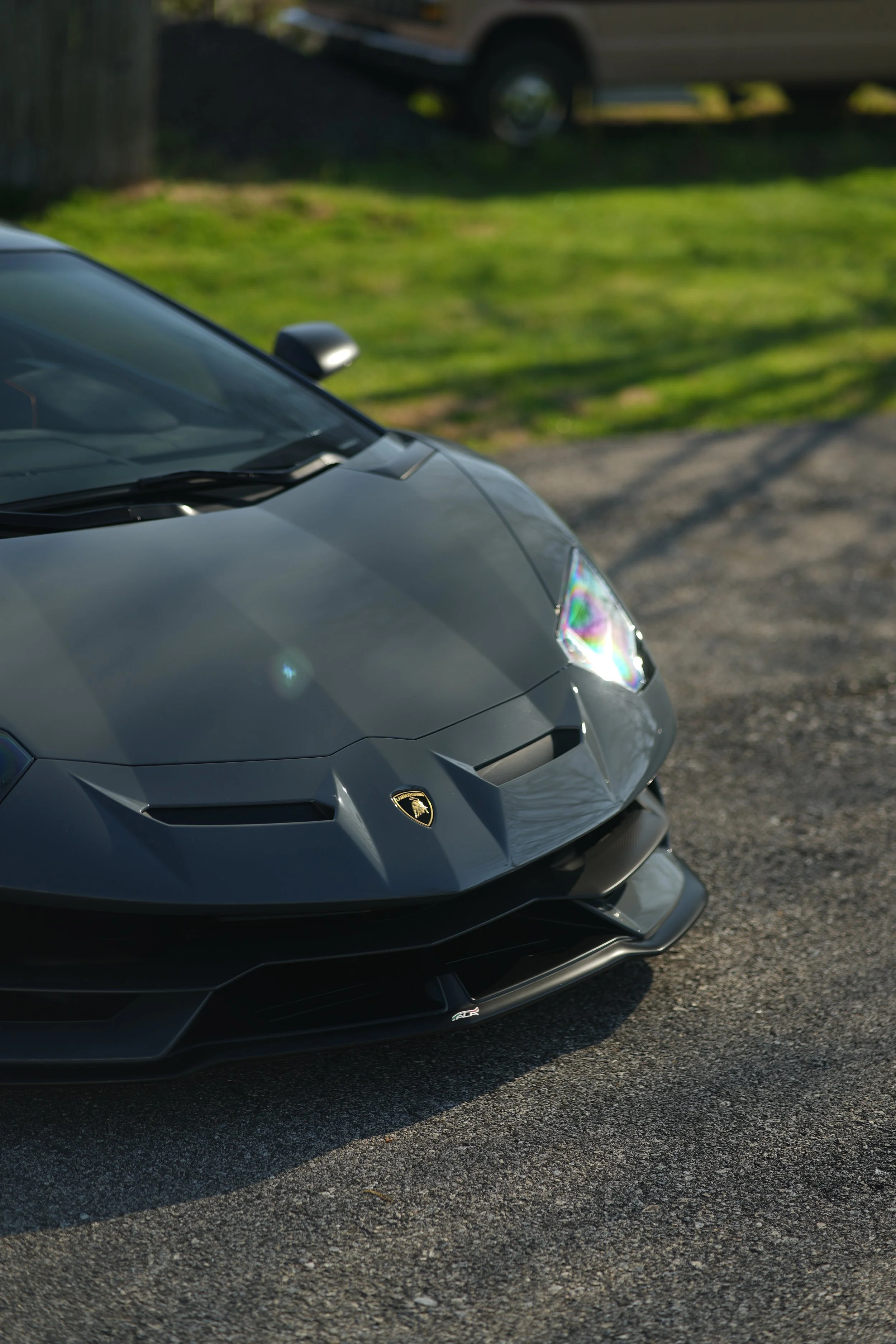 Close-up of a grey Lamborghini sports car parked on a gravel driveway with a grassy lawn and a vehicle in the background.