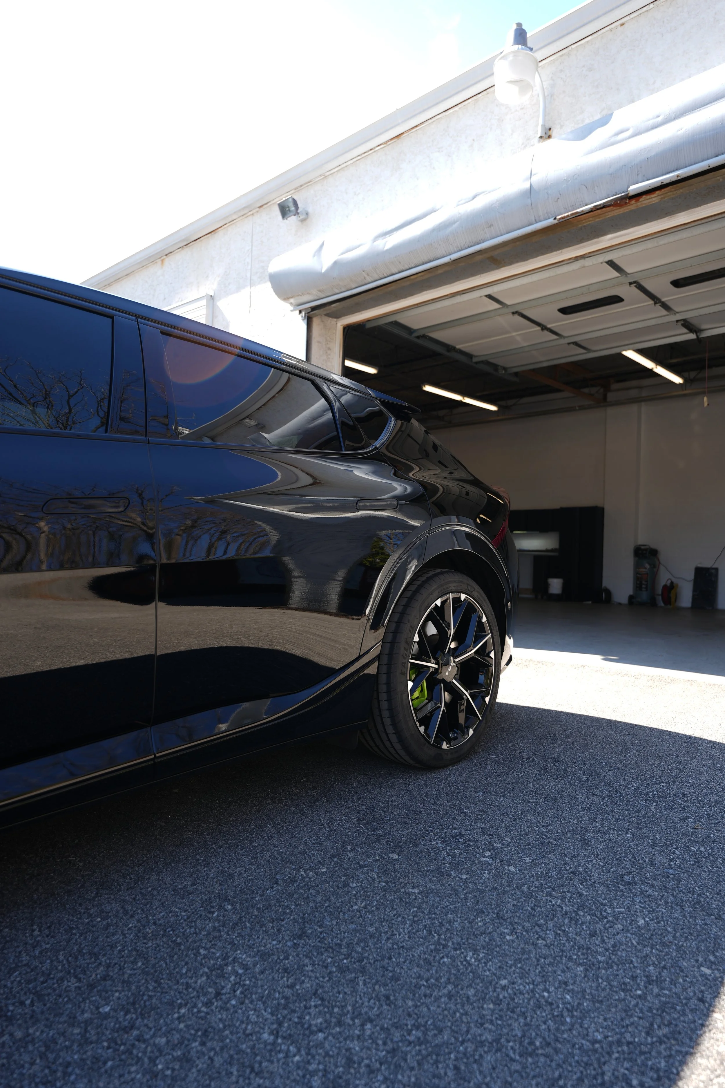 A black car parked in front of a garage with the garage door open. The garage has a white exterior with a slightly rusted auto-opened garage door and a white light fixture above it. The car's windows reflect the blue sky and nearby trees.