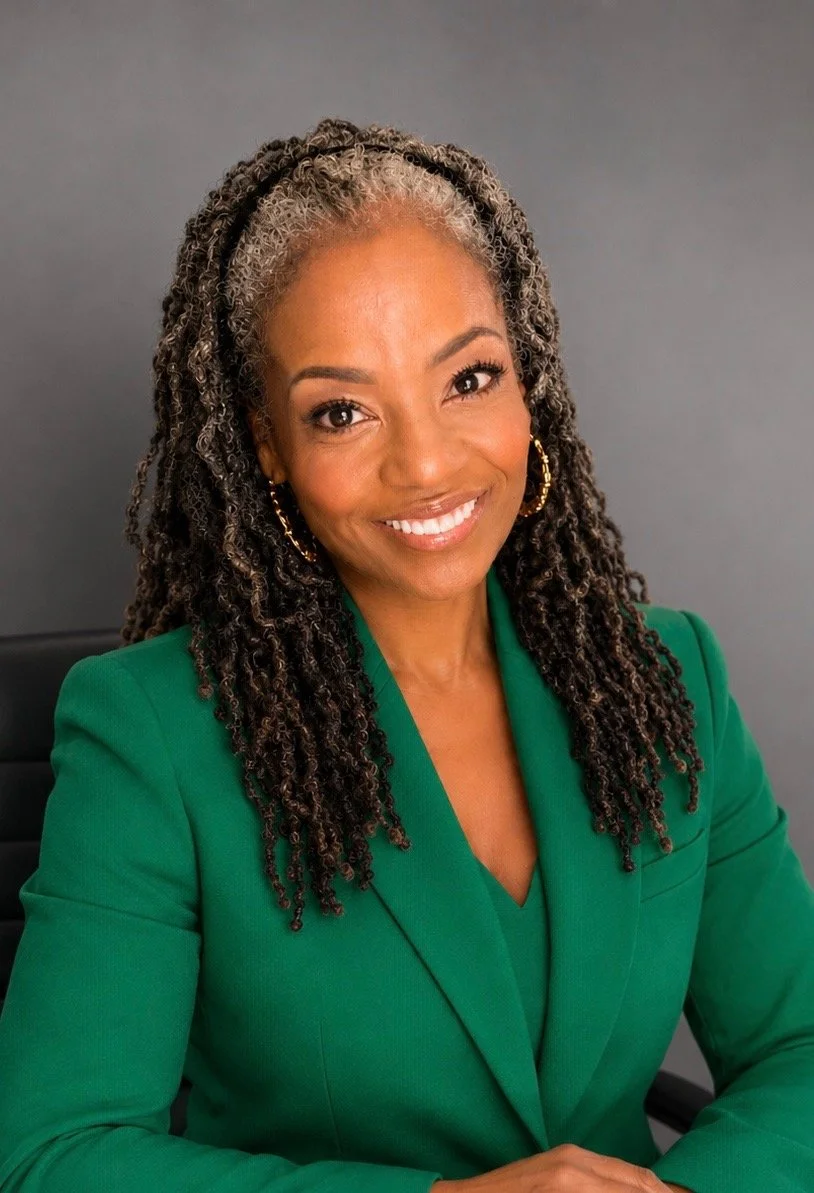Michelle Anderson wearing a green blazer, gold hoop earrings, and smiling, sitting against a gray background.